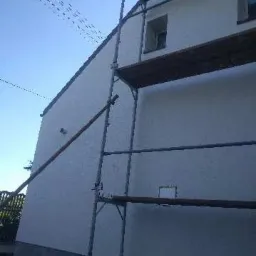 Ściana budynku w trakcie prac elewacyjnych, widoczne rusztowanie i świeżo nałożony tynk strukturalny. Dolna część ściany pomalowana na szaro.