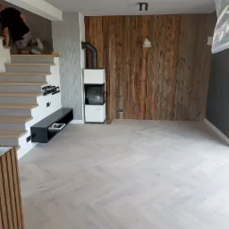 Jasnoszara podłoga ułożona w jodełkę francuską, drewniana ściana z desek, biały kominek z czarnym wkładem, czarna szafka RTV pod schodami z drewnianymi stopniami, betonowy sufit z czarnymi lampami.