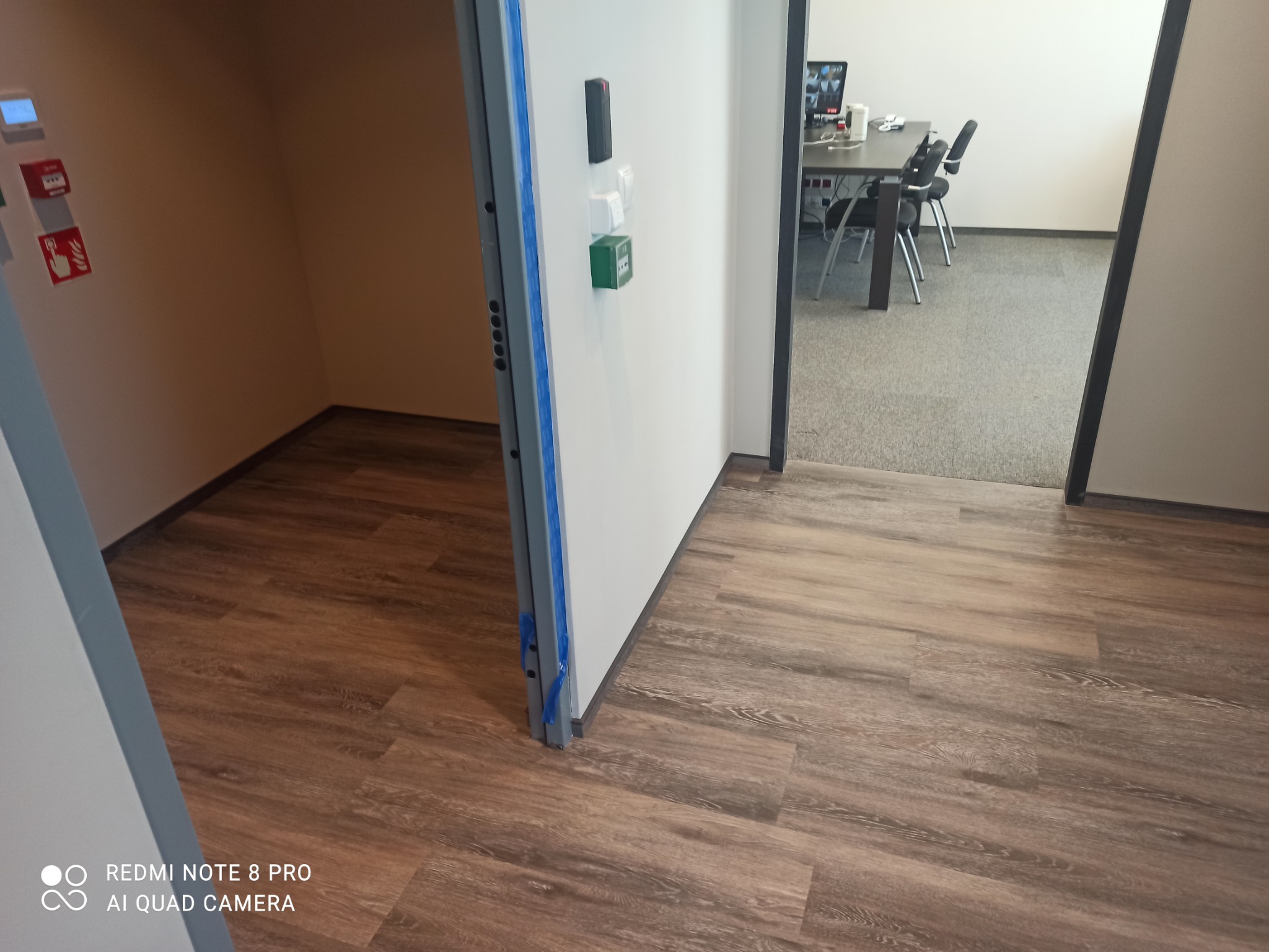 Nowa podłoga z paneli imitujących drewno w jasnym biurze, widok z korytarza na salę konferencyjną z szarym dywanem i stołem z krzesłami.