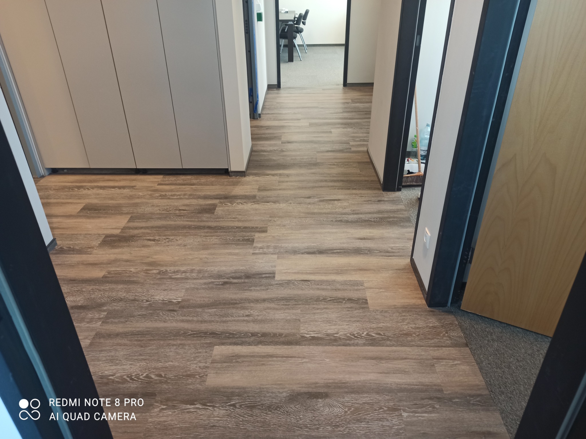 Ułożone panele podłogowe w biurowym korytarzu, widok z perspektywy wejścia, z widocznymi drzwiami i szafami po bokach.