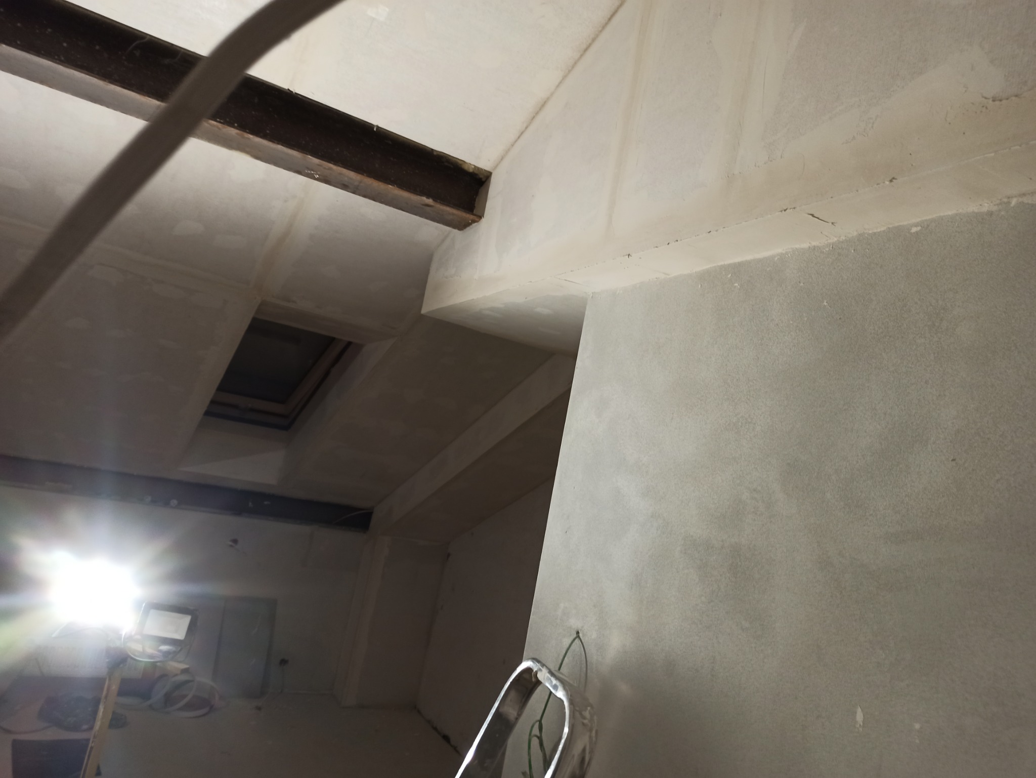 Surowe wnętrze w trakcie prac wykończeniowych, widoczna surowa ściana z nałożoną gładzią, metalowa belka konstrukcyjna pod sufitem, okno dachowe i drabina w tle, oświetlone mocnym światłem...