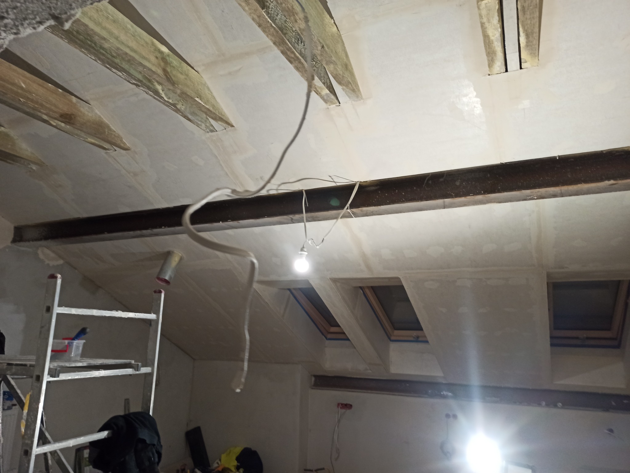 Skośny sufit poddasza w trakcie wykańczania płytami karton gips, widoczne drewniane elementy konstrukcyjne, stalowa belka nośna, okna dachowe i prowizoryczne oświetlenie na przewodach.