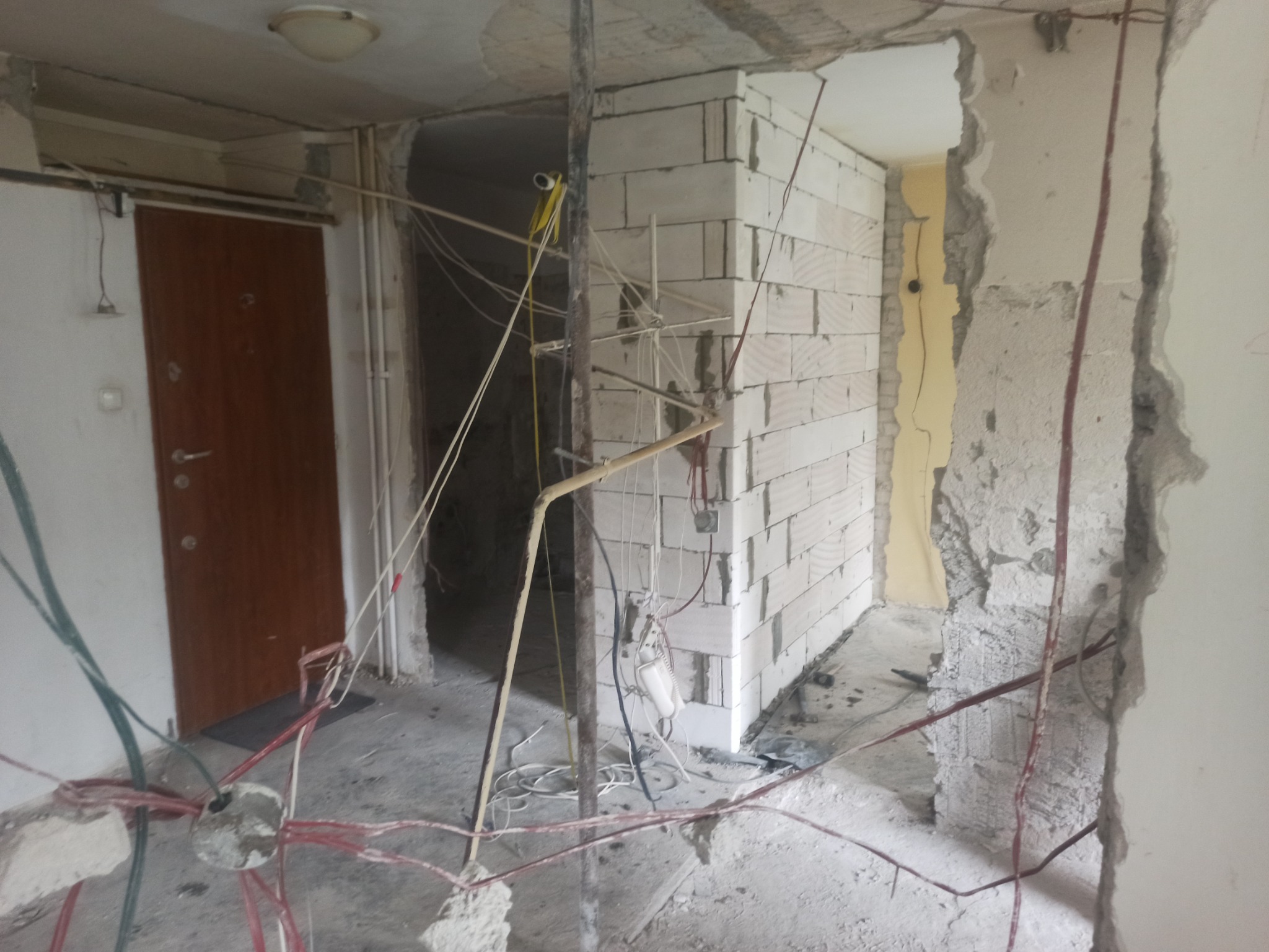 Wnętrze w trakcie przebudowy: widoczne surowe ściany, nowa ściana z bloczków, wystające kable elektryczne i metalowe podpory.