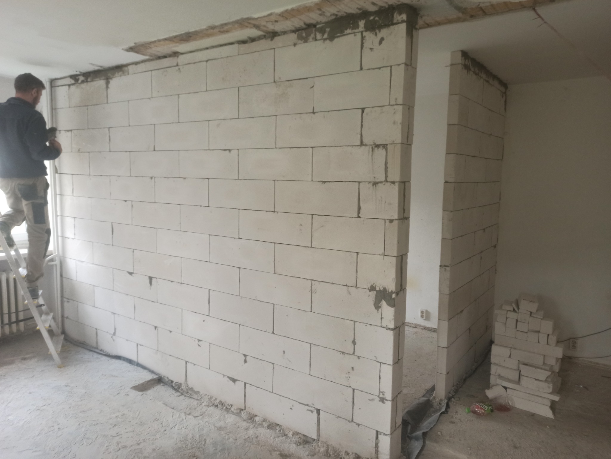 Wznoszenie ściany działowej z bloczków gazobetonowych, widoczny pracownik na drabinie, świeżo wymurowana ściana z widocznymi spoinami, stos bloczków obok otworu drzwiowego.