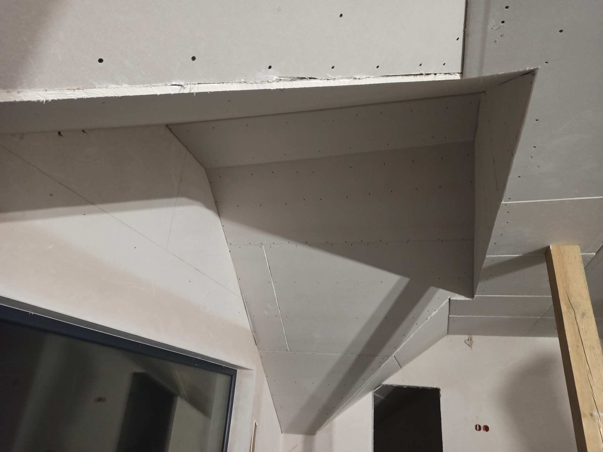 Sufit podwieszany z płyty gipsowo-kartonowej w trakcie montażu, widoczne ślady po wkrętach i łączenia płyt, drewniany wspornik tymczasowy, okno w dolnej części kadru.