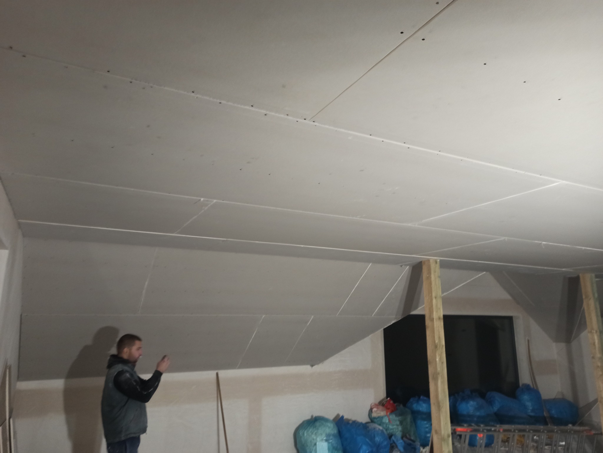 Montaż płyt kartonowo-gipsowych na skośnym suficie poddasza, widoczne łączenia płyt i tymczasowe drewniane podpory, pracownik sprawdza postęp prac.