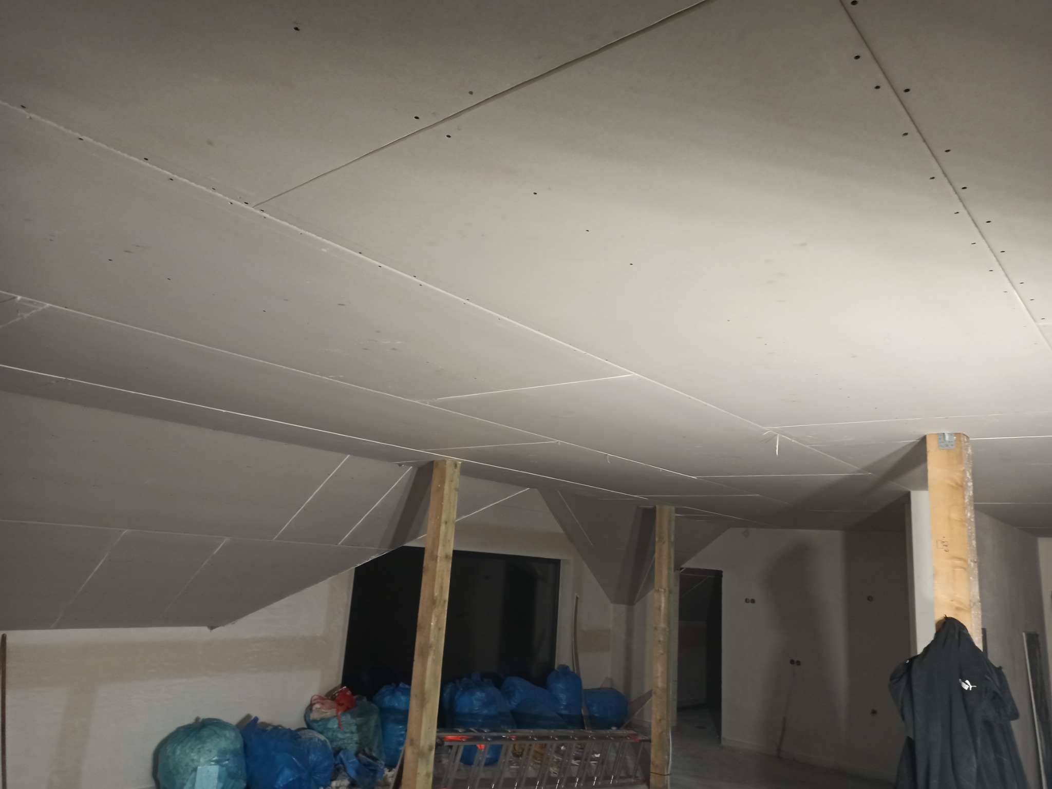 Montaż płyt kartonowo-gipsowych na skośnym suficie poddasza, widoczne drewniane podpory i worki z odpadami budowlanymi.