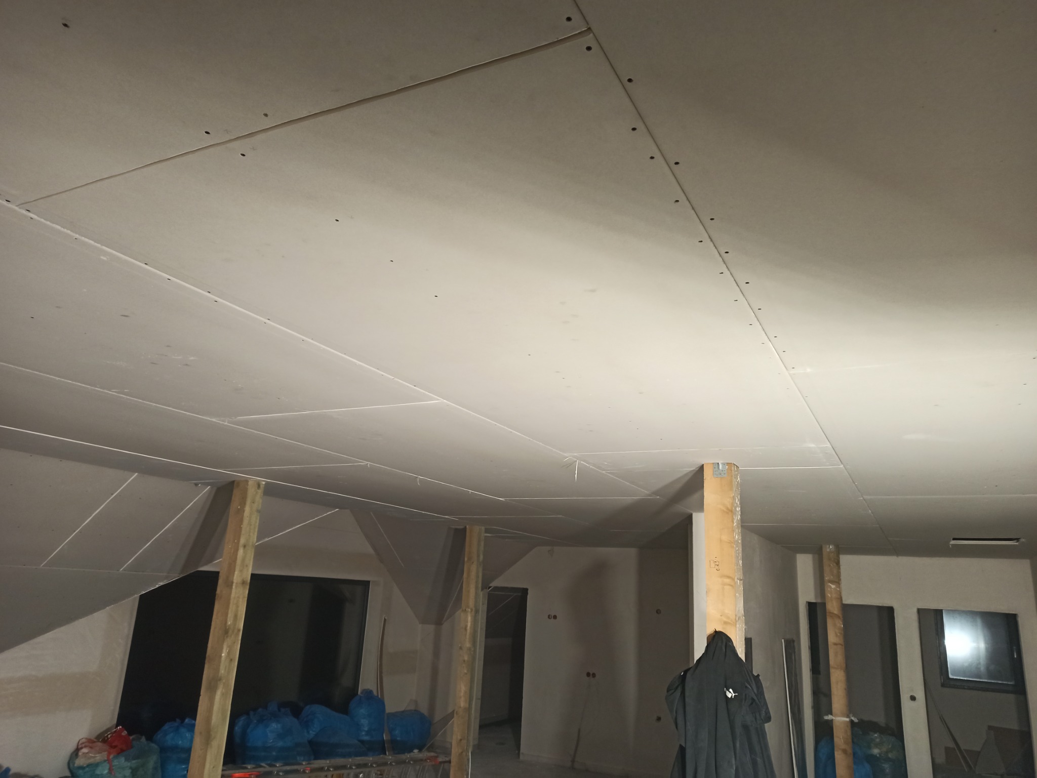 Montaż sufitu podwieszanego z płyt kartonowo-gipsowych w pomieszczeniu ze skośnym dachem, widoczne drewniane podpory i niebieskie worki z materiałami budowlanymi.