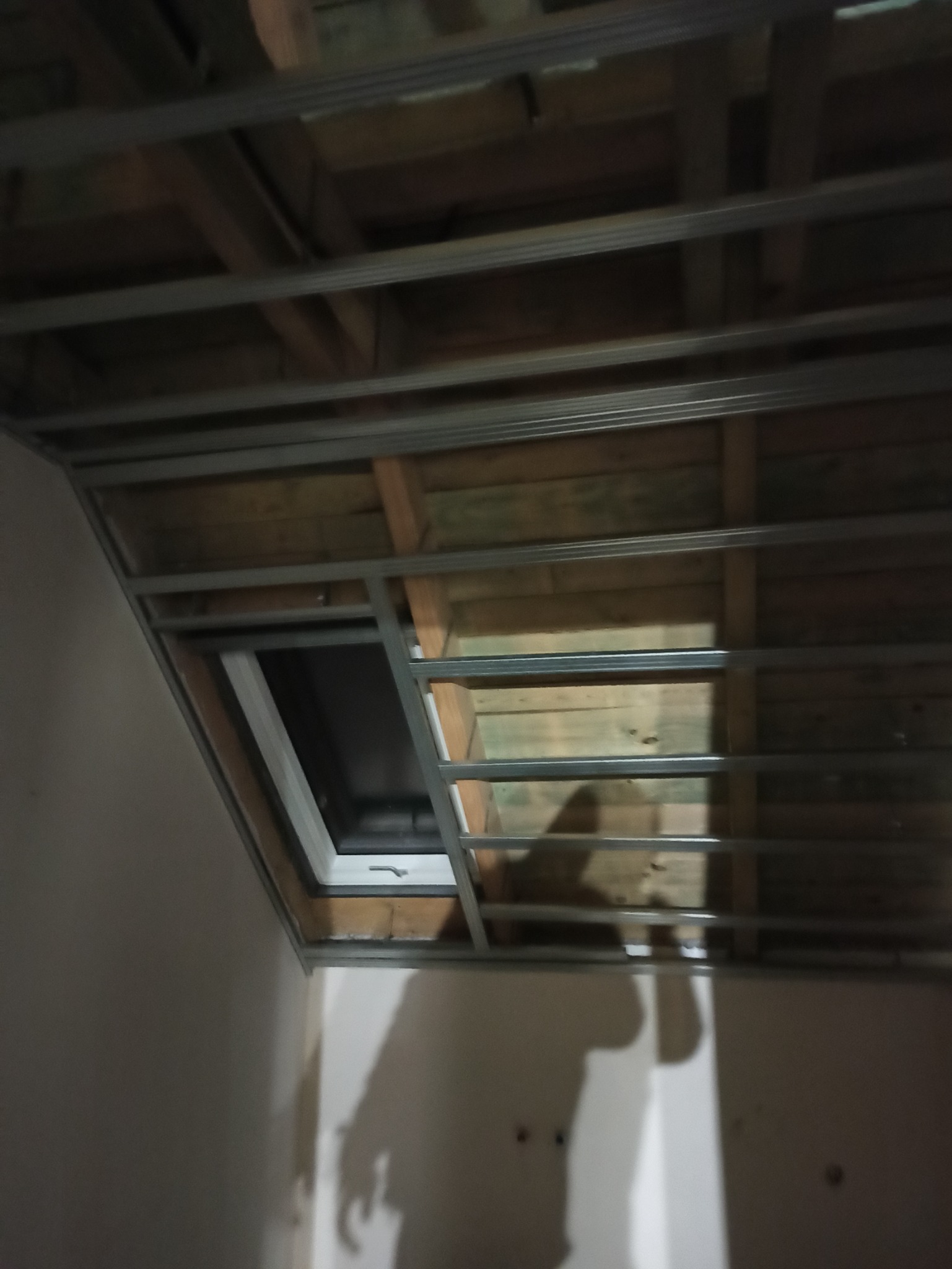 Stelaż z profili metalowych pod zabudowę sufitu z widoczną drewnianą konstrukcją stropu i fragmentem okna dachowego.