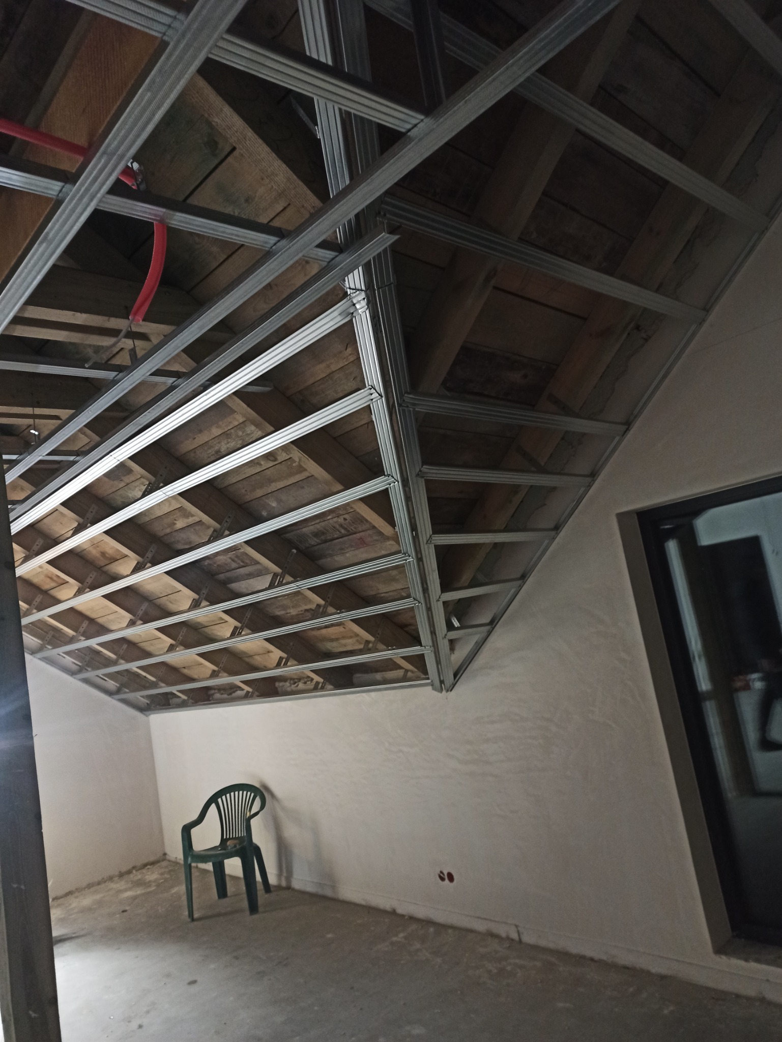 Metalowa konstrukcja sufitu podwieszanego z widoczną starą drewnianą konstrukcją dachu, czerwony przewód instalacyjny i zielone krzesło w pustym pomieszczeniu z oknem.