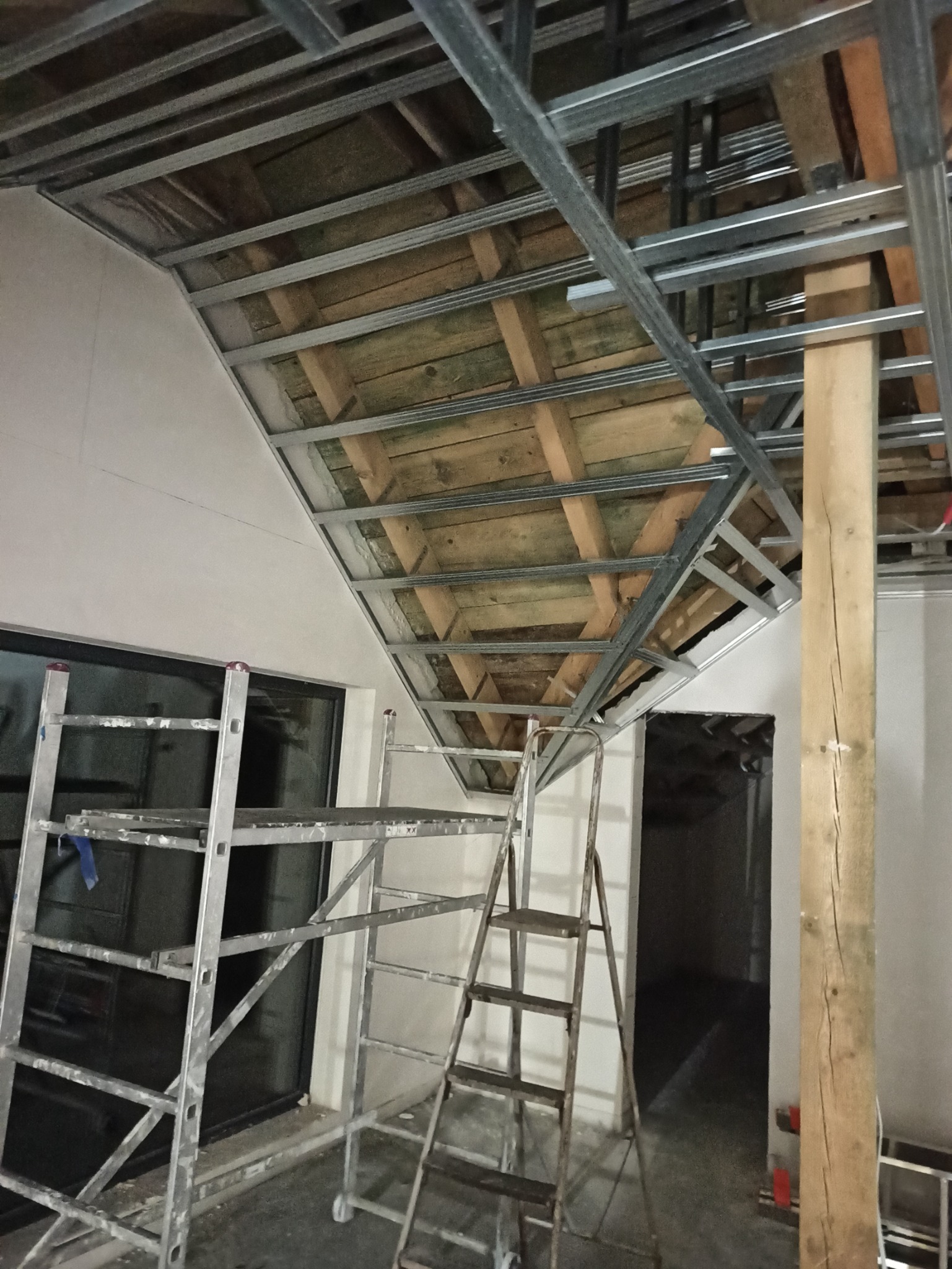 Stelaż z profili metalowych pod płytę karton-gips na skośnym poddaszu, widoczne drewniane belki stropu, drabiny i słup podtrzymujący konstrukcję.