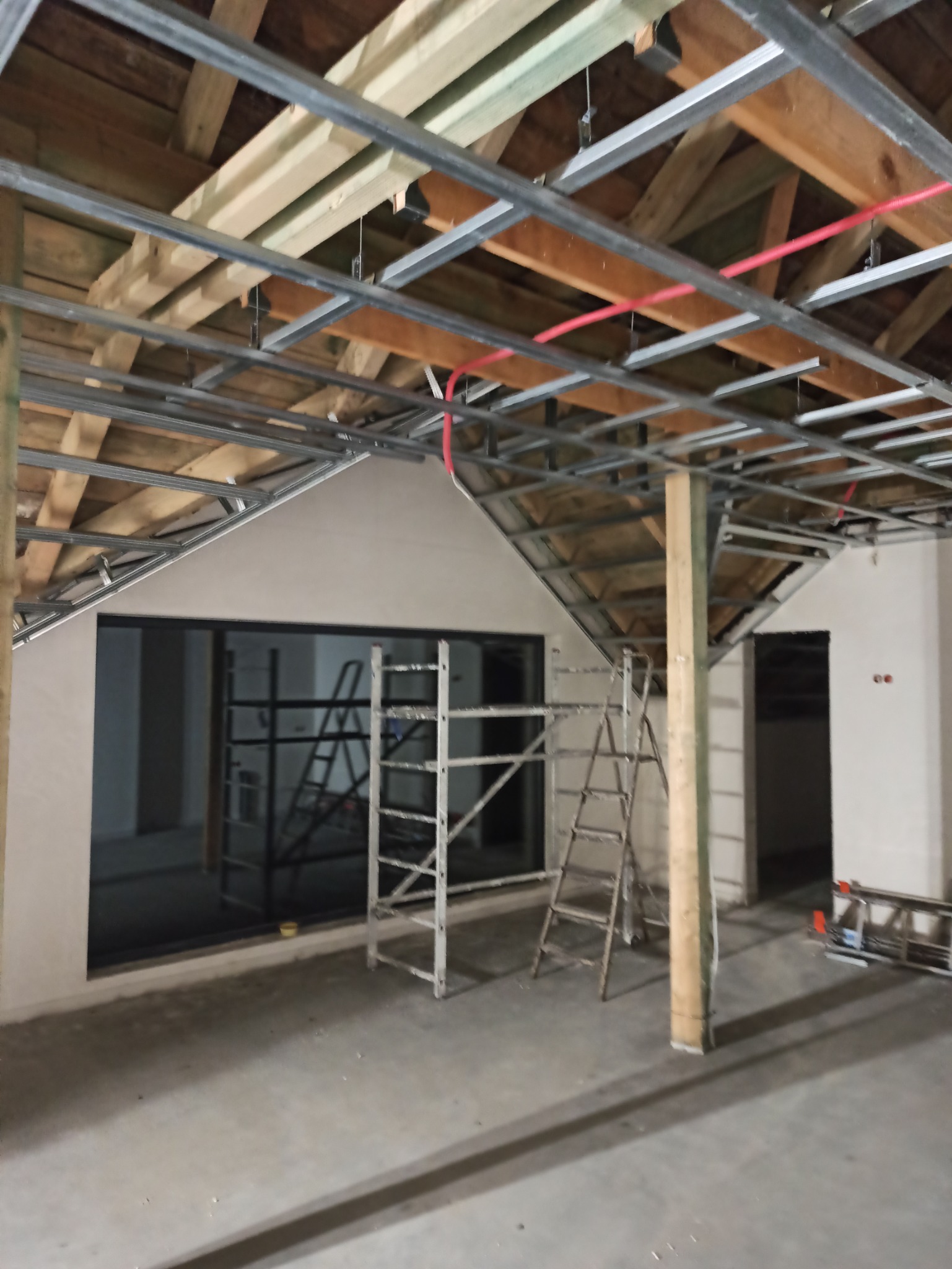 Wnętrze w trakcie prac remontowych, widoczne metalowe profile sufitowe pod drewnianą konstrukcją dachu, ściany przygotowane pod malowanie, okno z widocznym rusztowaniem oraz drabiny.