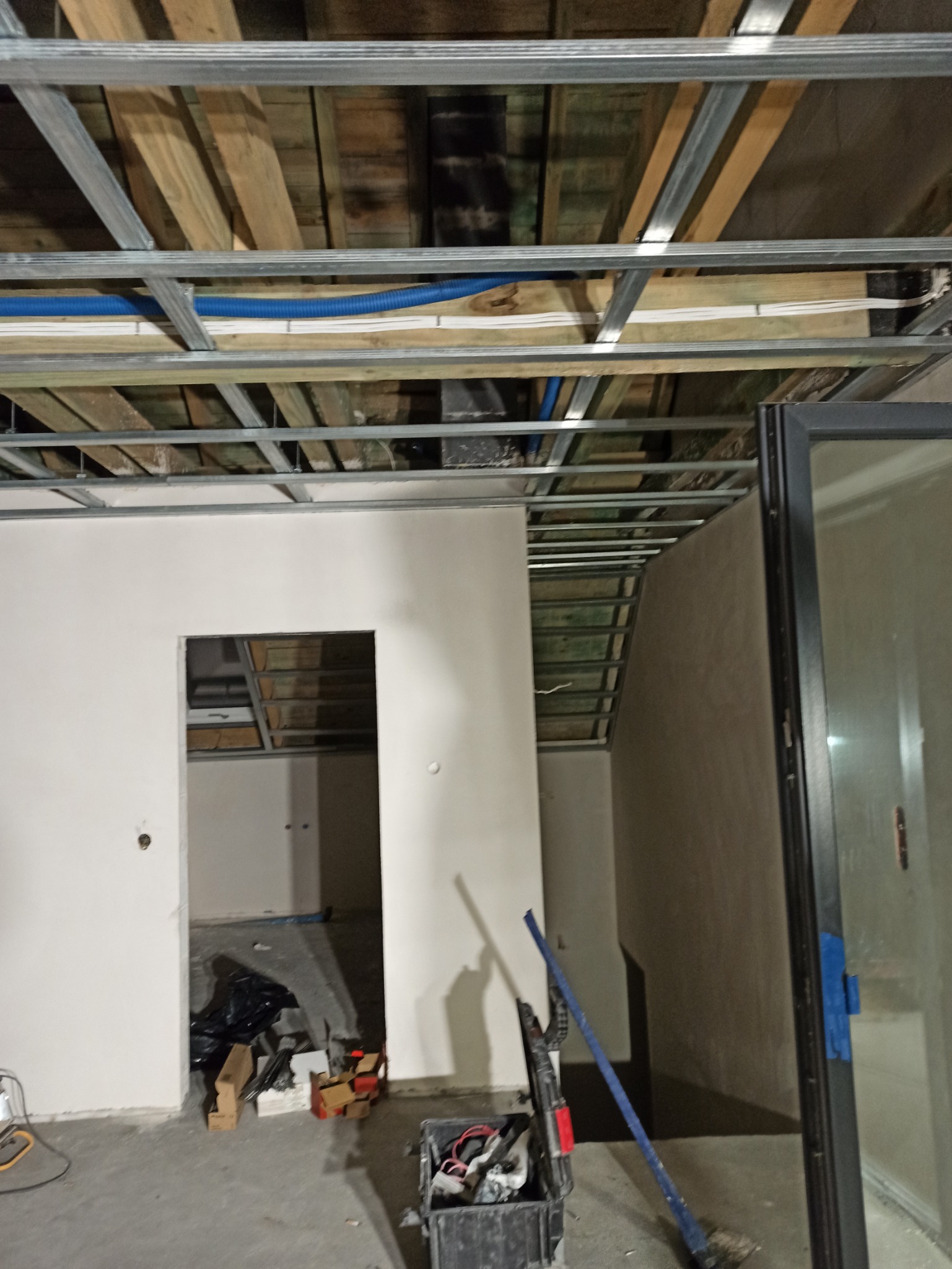 Wnętrze w trakcie prac wykończeniowych: widoczny szkielet sufitu podwieszanego z instalacją, surowe ściany z otworami drzwiowymi oraz narzędzia w skrzynce na podłodze.