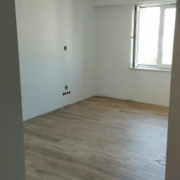 Pokój po położeniu paneli podłogowych, widoczne niedokończone ściany, wystające kable z sufitu i narzędzia na podłodze.