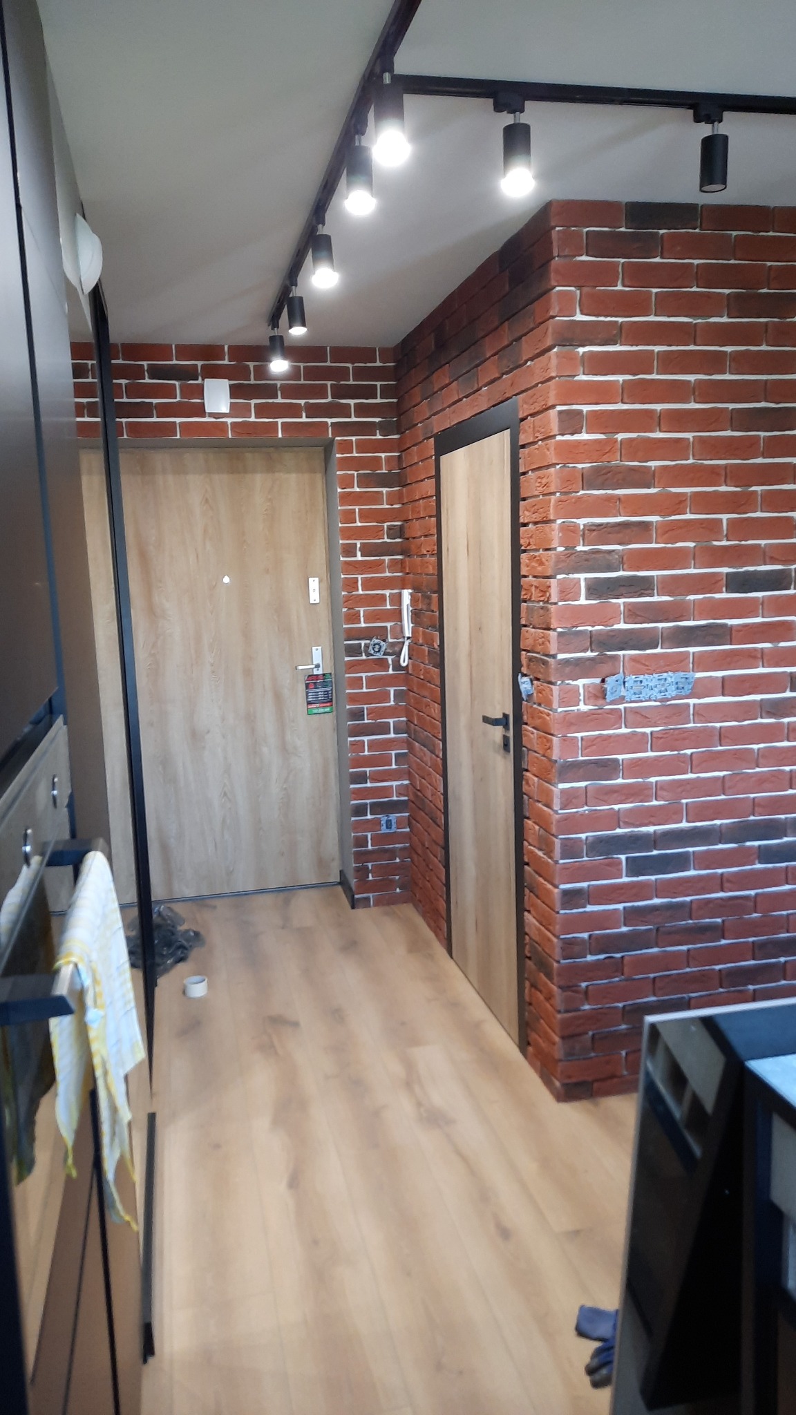 Korytarz wykończony czerwoną cegłą, oświetlony reflektorami na szynie, z dwojgiem drzwi o jasnym, drewnianym wykończeniu i panelami podłogowymi w ciepłym odcieniu drewna.