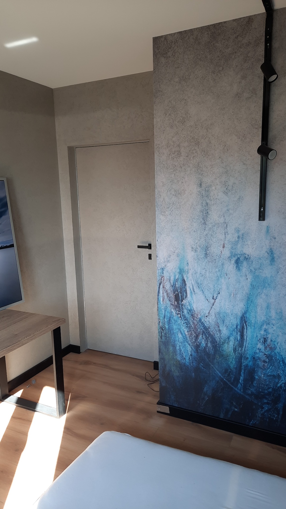 Pokój po odświeżeniu: ściana z efektem dekoracyjnym w odcieniach szarości i błękitu, minimalistyczne oświetlenie punktowe na szynie, jasne panele podłogowe i fragment mebla z drewnianym blatem.