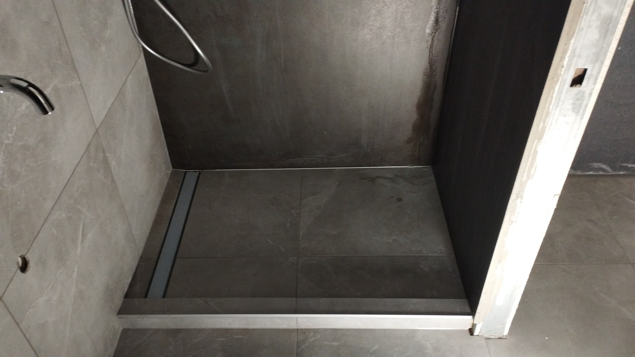 Nowoczesny brodzik z odpływem liniowym, wykończony szarą płytką imitującą kamień, fragment kabiny prysznicowej w trakcie prac wykończeniowych, widoczna surowa ściana i fragment ościeżnicy.