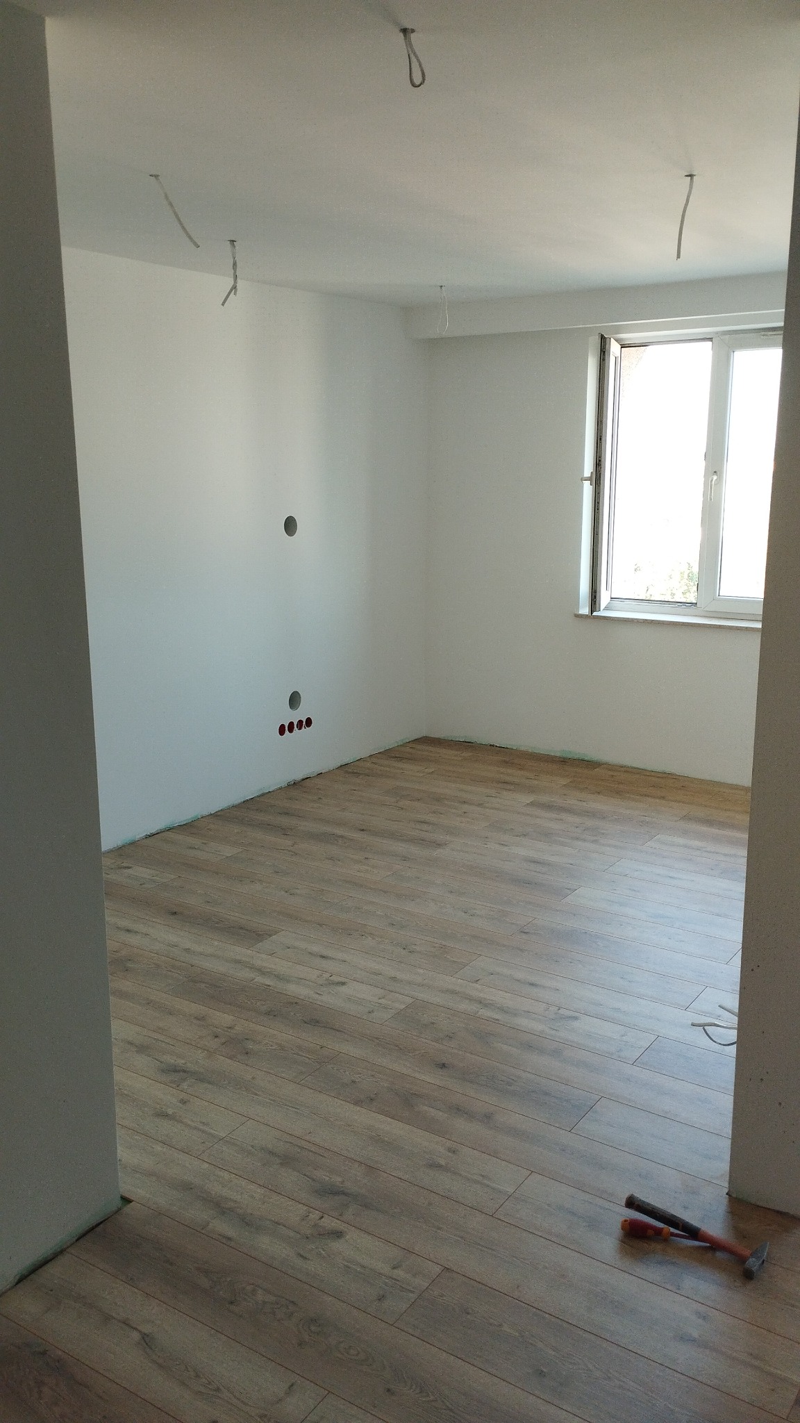 Pokój po położeniu paneli podłogowych, widoczne niedokończone ściany, wystające kable z sufitu i narzędzia na podłodze.