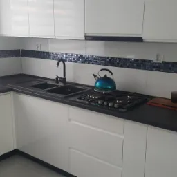 Nowoczesna kuchnia z białymi, lakierowanymi frontami, czarnym blatem, mozaiką na ścianie i niebieskim czajnikiem na płycie gazowej.