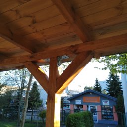 Czarny usługi budowlane - Drewniana konstrukcja z widocznym dachem, słupem i elementami mocującymi na tle budynku i zieleni.