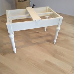 Czarny usługi budowlane - Biała, niedokończona rama meblowa z toczonymi nogami stoi na jasnej podłodze, widoczne elementy montażowe i kartonowe pudło w tle.