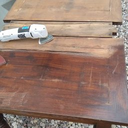 Czarny usługi budowlane - Renowacja drewnianego stołu: szlifierka oscylacyjna na blacie stołu, widoczne ślady szlifowania, obok drugi blat w trakcie obróbki, tło z kamyków.