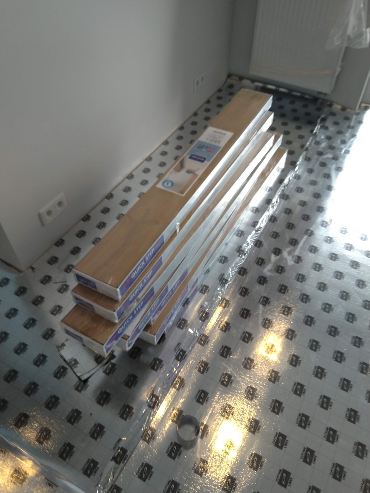 Stos paczek paneli podłogowych Quick Step leżący na podkładzie izolacyjnym z nadrukiem, przygotowany do montażu w rogu pokoju.