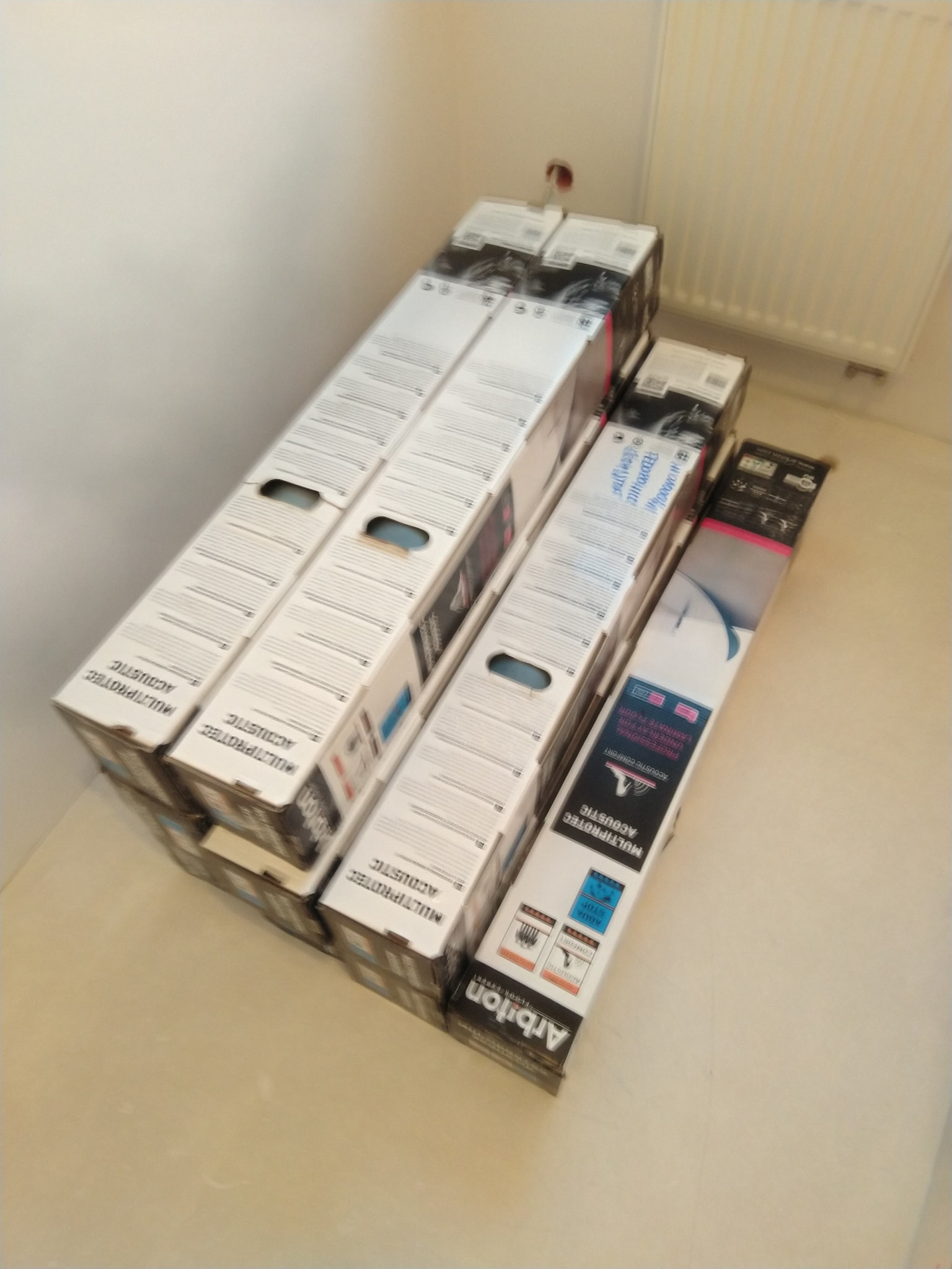 Paczki z podkładem akustycznym pod panele podłogowe, ułożone w stos na podłodze w rogu pomieszczenia przed montażem. Widoczne etykiety producenta Arbiton MULTIPROTEC ACOUSTIC.