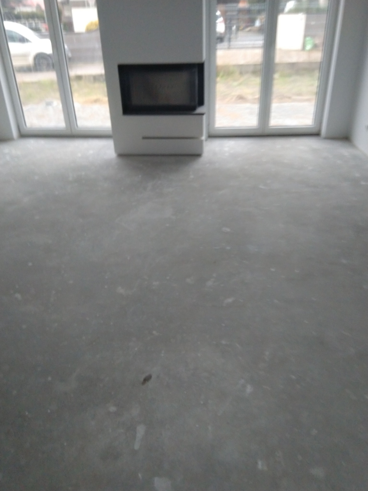 Przygotowane podłoże pod parkiet lub płytki w surowym stanie, widoczny kominek i okna balkonowe.