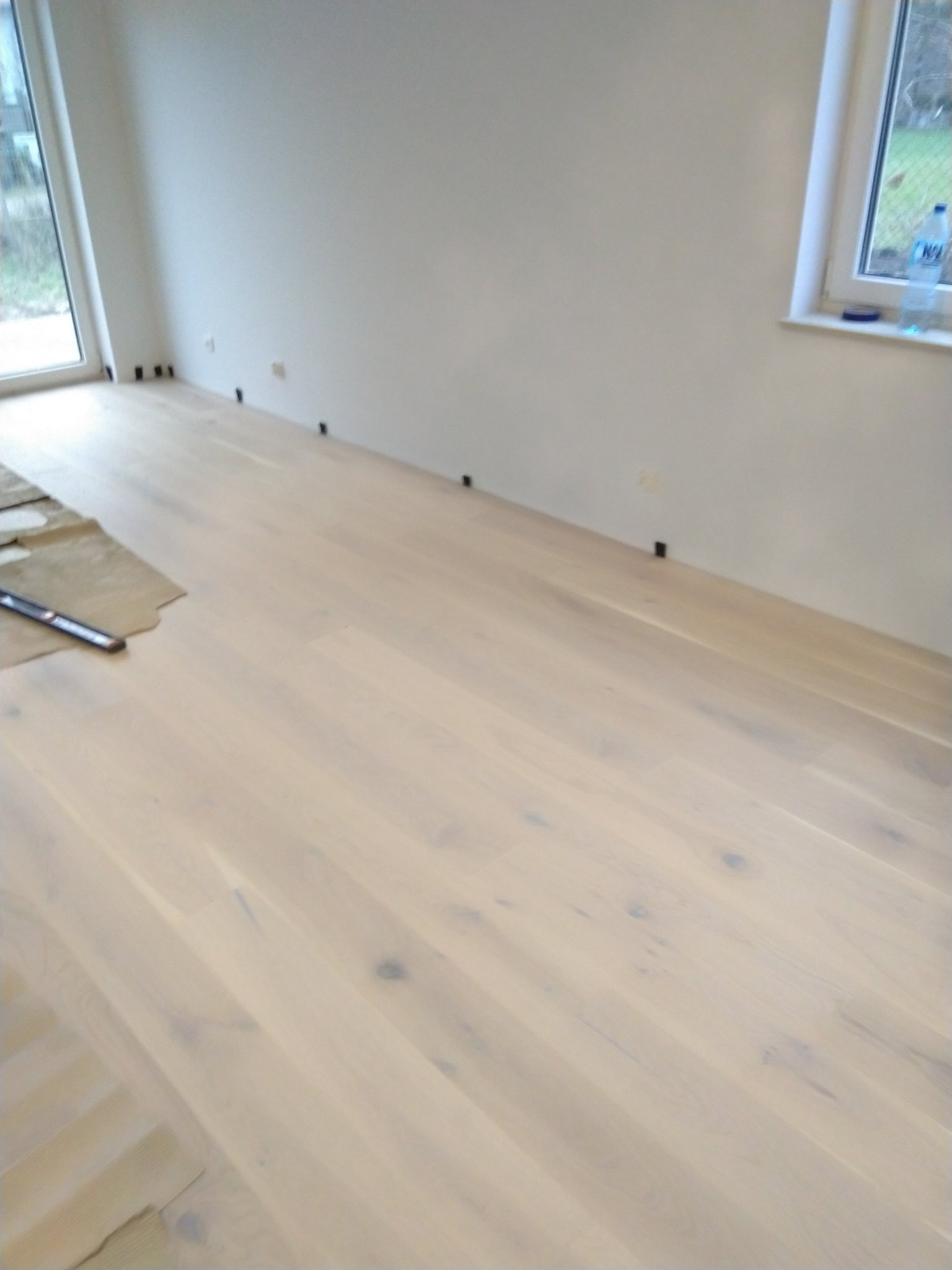 Jasny, świeżo ułożony parkiet w jodełkę, częściowo przykryty kartonem ochronnym i poziomnicą, widoczne listwy przypodłogowe w trakcie montażu.