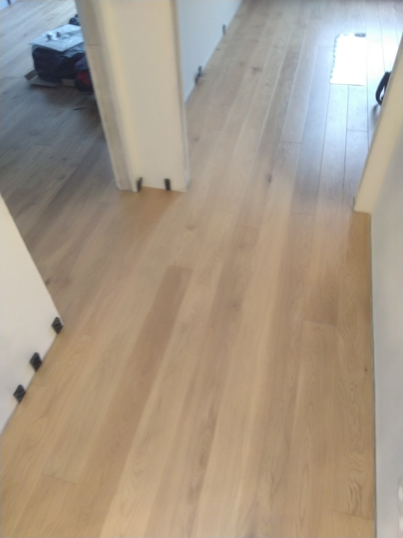 Jasne, drewniane deski podłogowe ułożone wzdłuż korytarza, widoczne fragmenty ścian z czarnymi elementami montażowymi przy podłodze, na podłodze w tle widoczne materiały budowlane zabezpieczone folią.