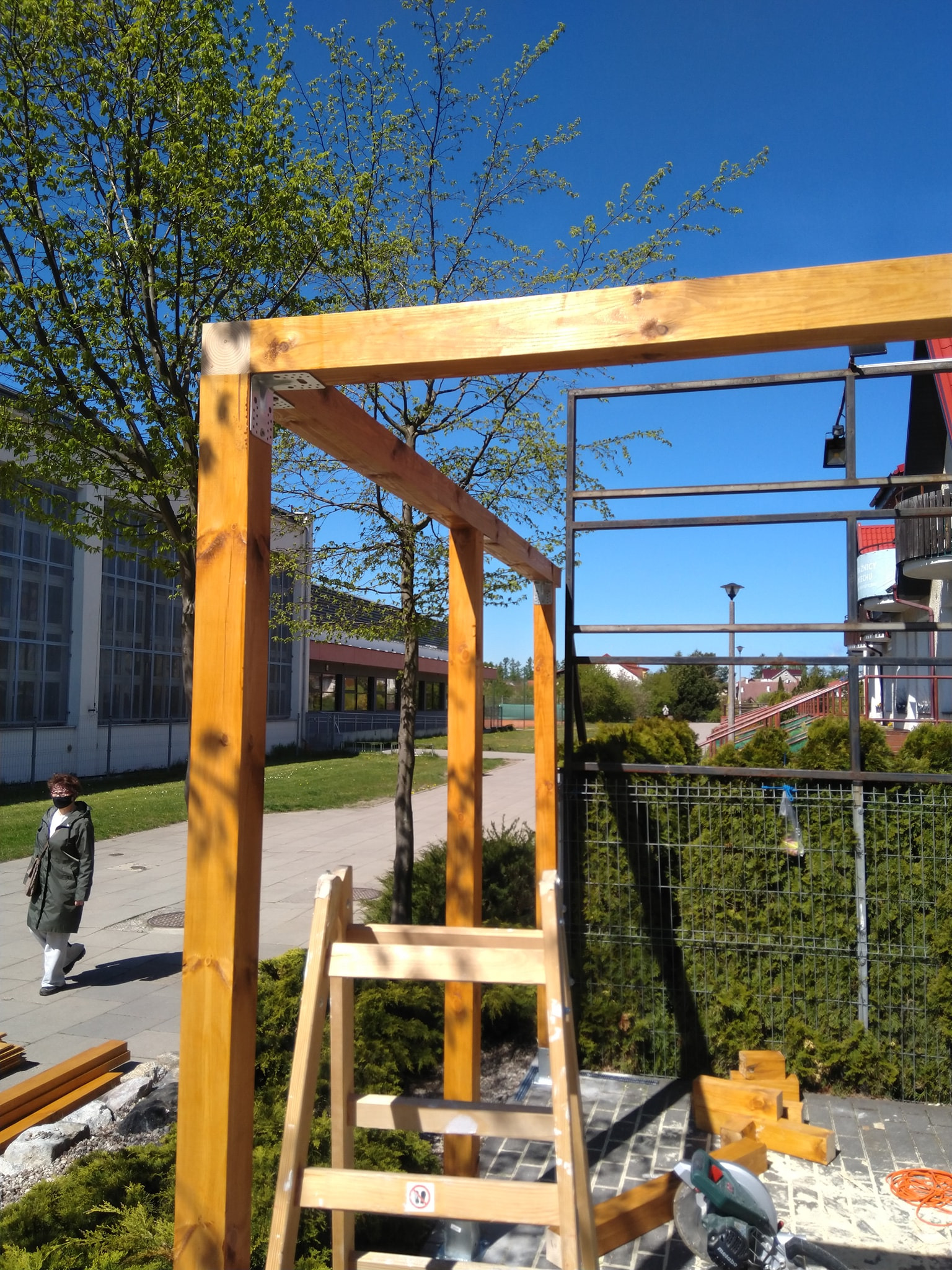 Konstrukcja drewniana altany w trakcie budowy, widoczne metalowe łączniki, drabina, piła elektryczna i chodząca osoba w tle.