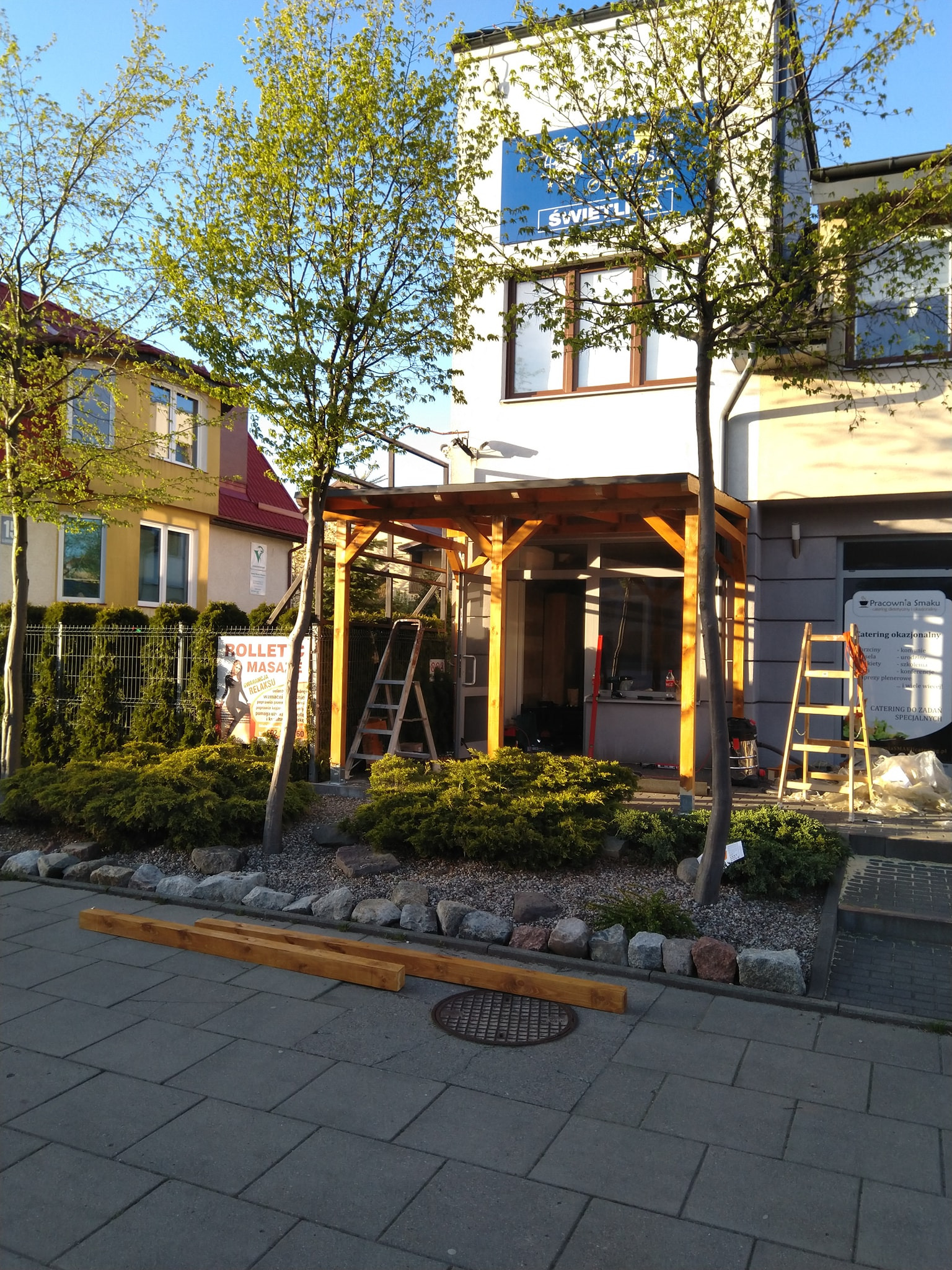 Drewniana konstrukcja zadaszenia tarasu w trakcie montażu przed budynkiem, widoczne elementy konstrukcyjne, drabina i narzędzia.