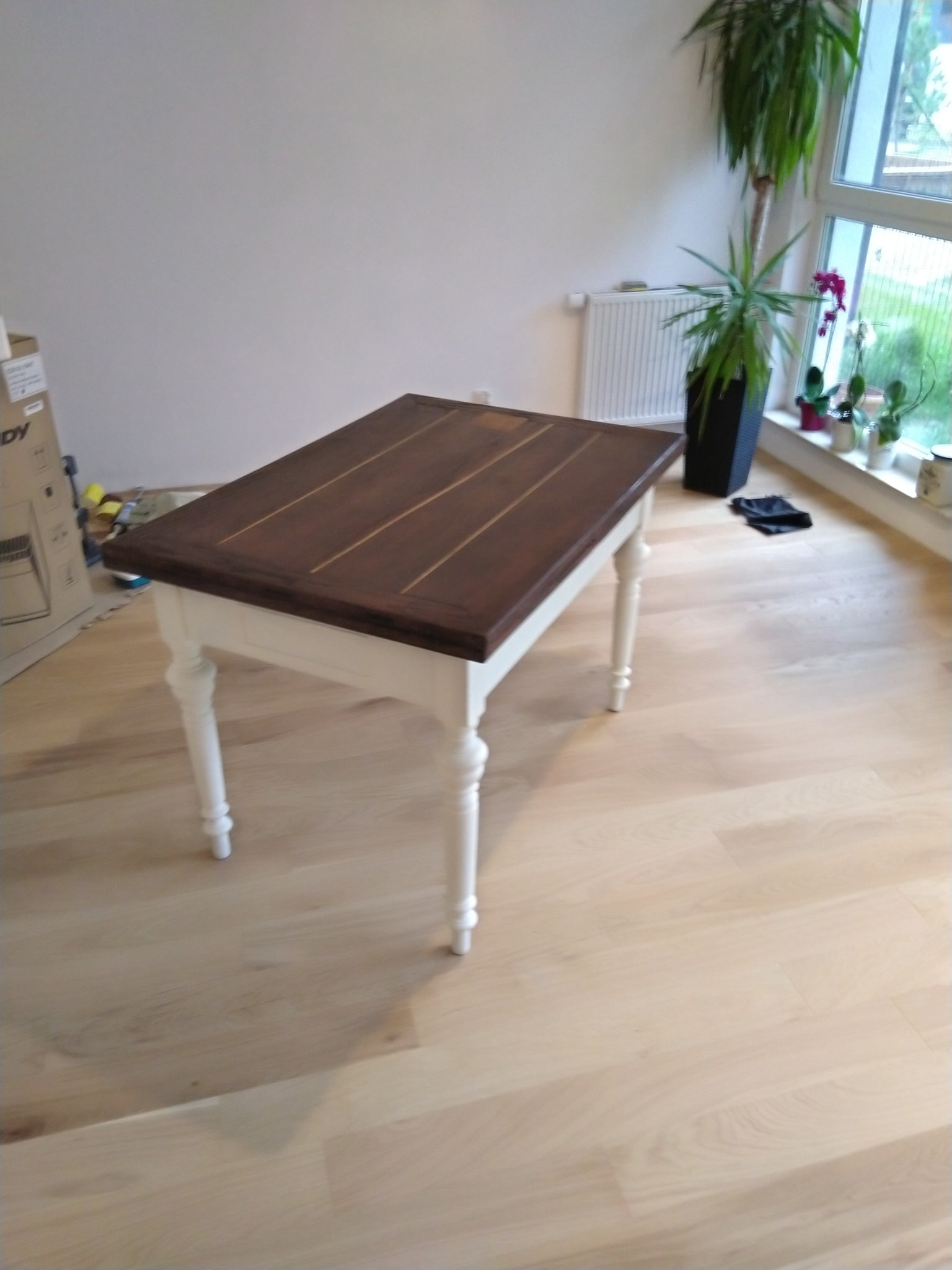 Stół z ciemnobrązowym blatem i białymi, toczonymi nogami na jasnej drewnianej podłodze, w tle biała ściana, grzejnik i roślina doniczkowa przy oknie.