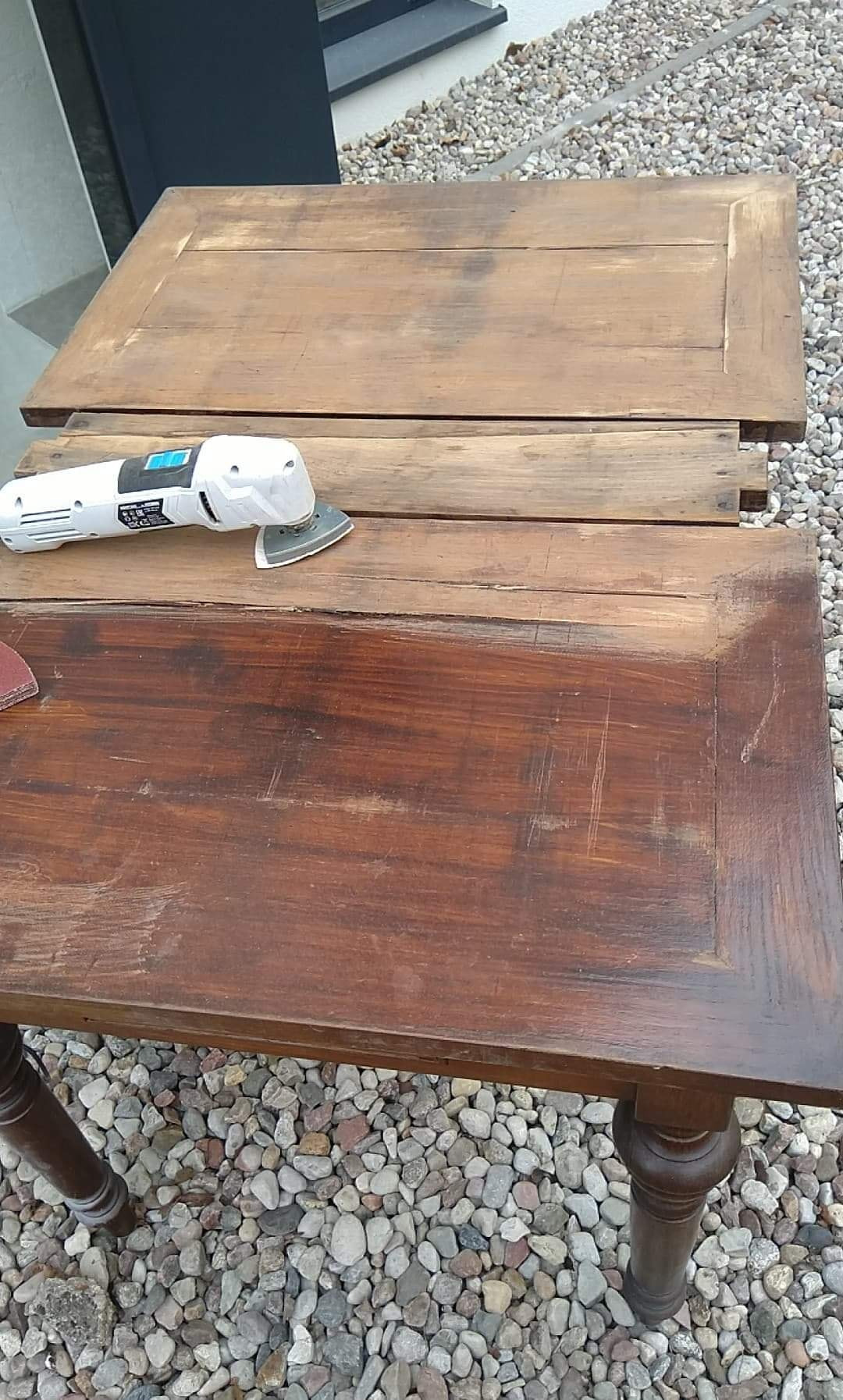Renowacja drewnianego stołu: szlifierka oscylacyjna na blacie stołu, widoczne ślady szlifowania, obok drugi blat w trakcie obróbki, tło z kamyków.