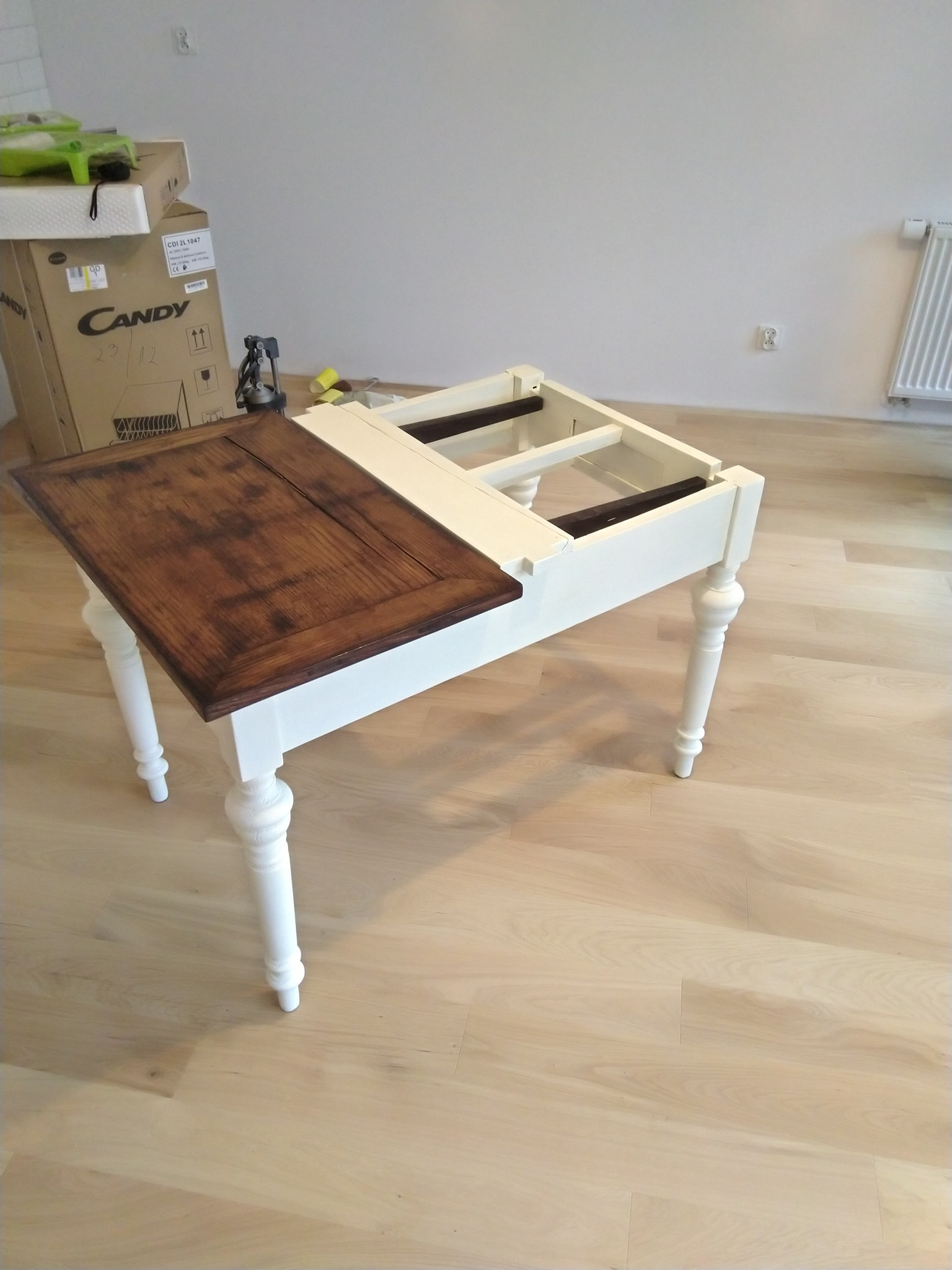 Stół z blatem w odcieniach brązu i białymi nogami w trakcie montażu, widoczna konstrukcja bez pełnego blatu, karton w tle.