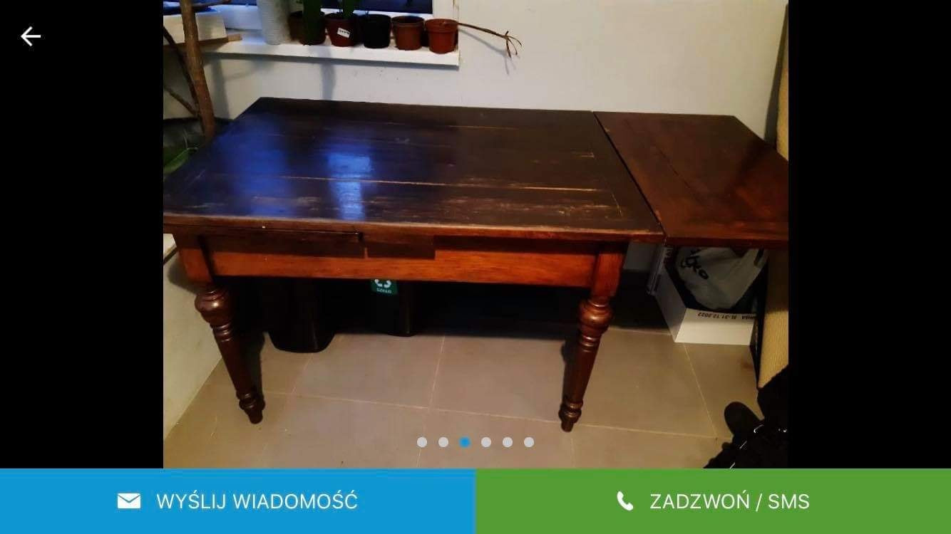 Stary, drewniany stół z wysuwanym blatem i toczonymi nogami, widoczne ślady użytkowania i odnowiona powierzchnia, na tle ściany z parapetem.