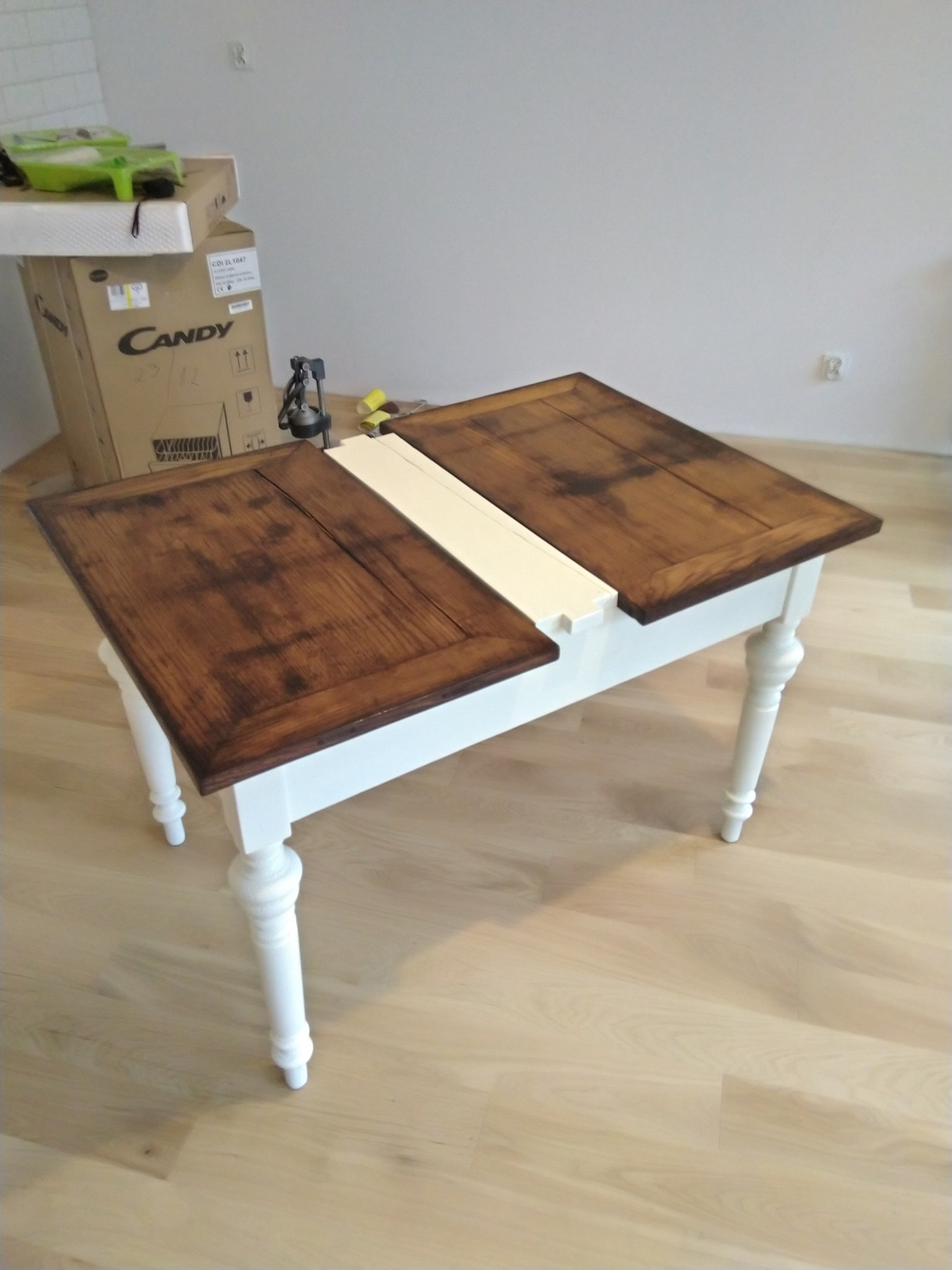 Stół z dwiema drewnianymi deskami blatu w odcieniach brązu i białym elementem łączącym, na tle kartonu ze sprzętem AGD i jasnej podłogi.