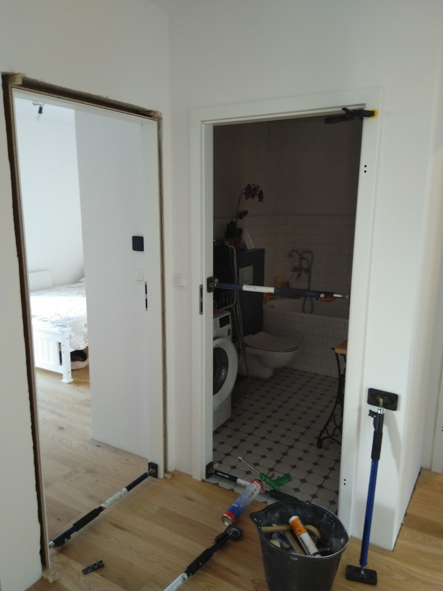 Montaż ościeżnic drzwiowych w dwóch otworach, widoczne rozpórki i narzędzia, w tle łazienka i sypialnia.