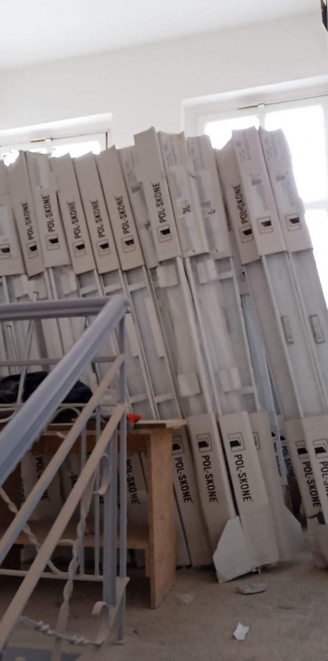 Paczki z białymi drzwiami marki POL-SKONE, opierające się o ścianę w jasnym pomieszczeniu w trakcie remontu, widoczne metalowe schody i drewniany stołek.