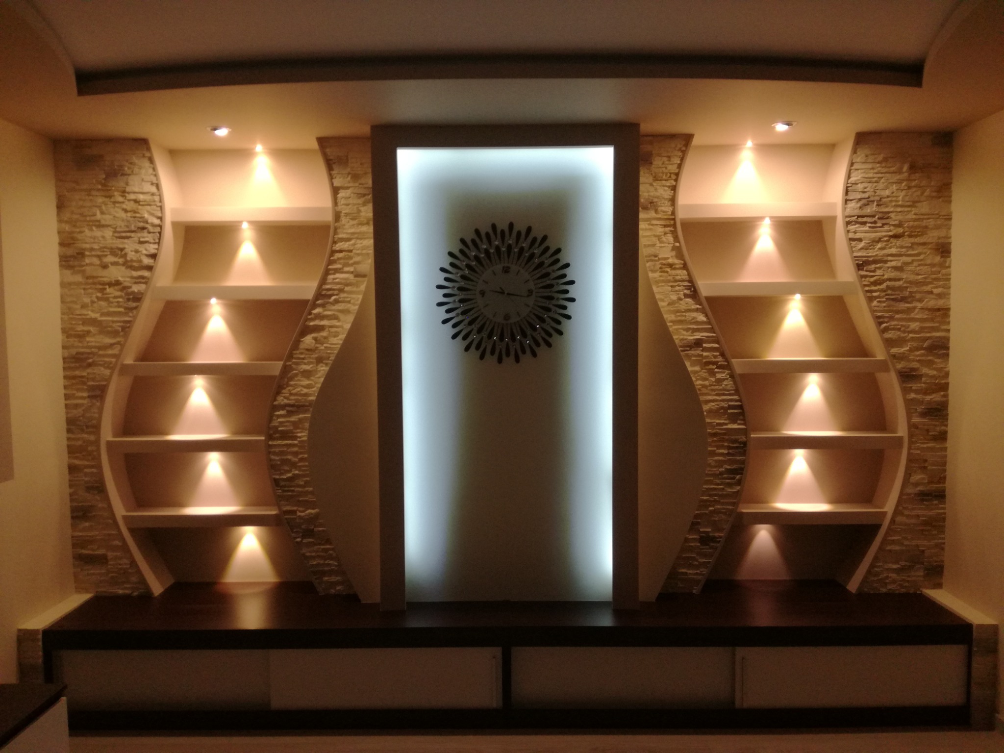 Oświetlona ściana z dekoracyjną okładziną z kamienia i półkami o falistym kształcie, centralnie umieszczony podświetlany panel z zegarem w stylu słońca.