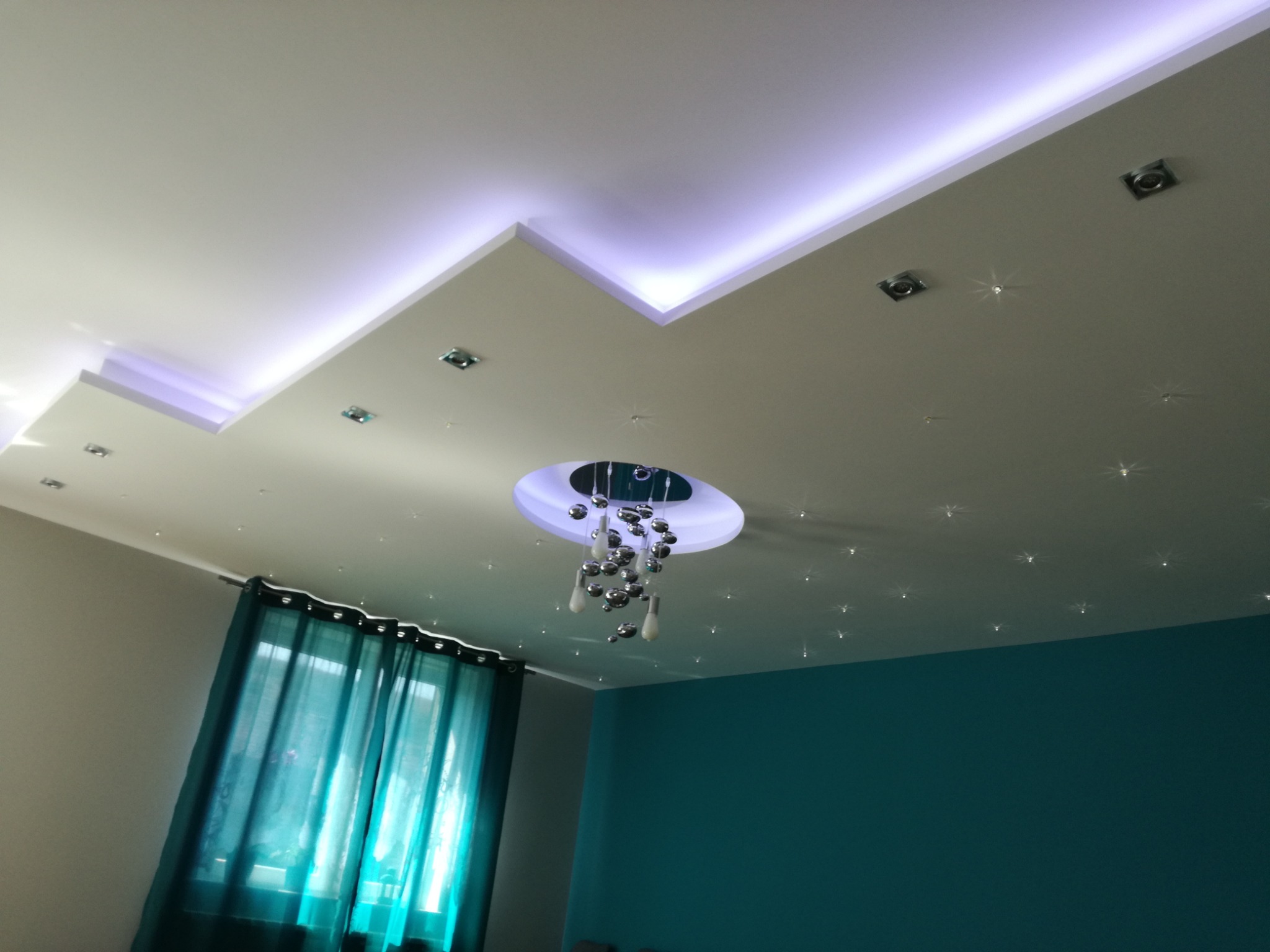 Sufit z podwieszanym elementem z oświetleniem LED, dekoracyjnymi punktami świetlnymi imitującymi gwiazdy oraz nowoczesnym żyrandolem z chromowanymi kulami, widok z dołu.