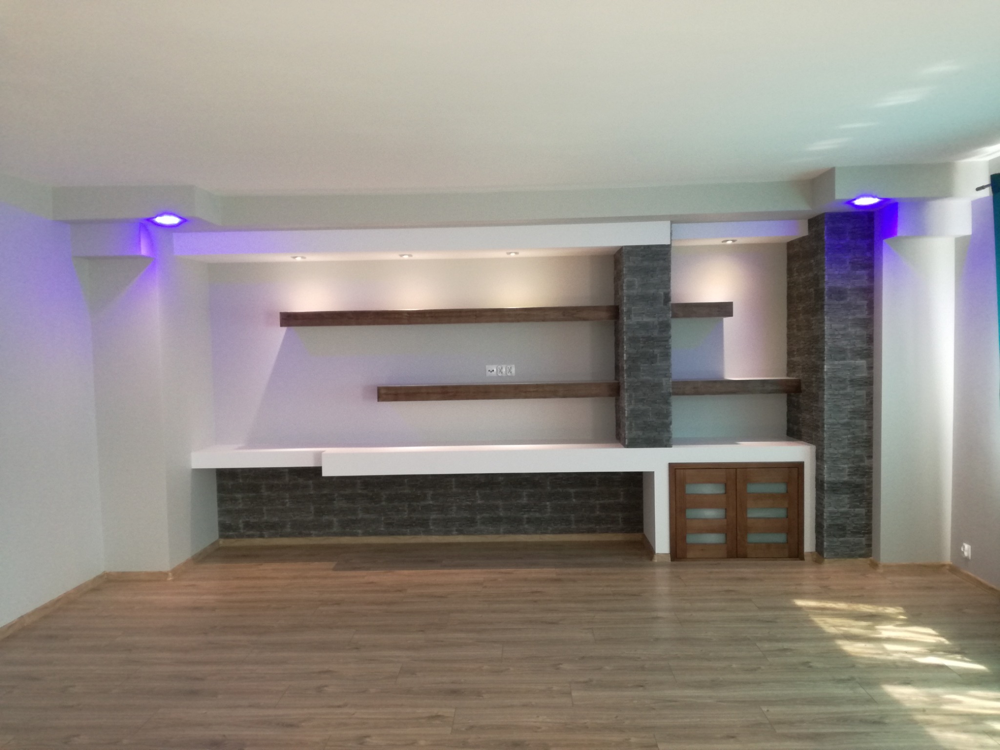 Nowoczesny salon po remoncie z drewnianą podłogą, półkami i dekoracyjnym kamieniem na ścianie, oświetlony światłem LED w kolorze fioletowym.