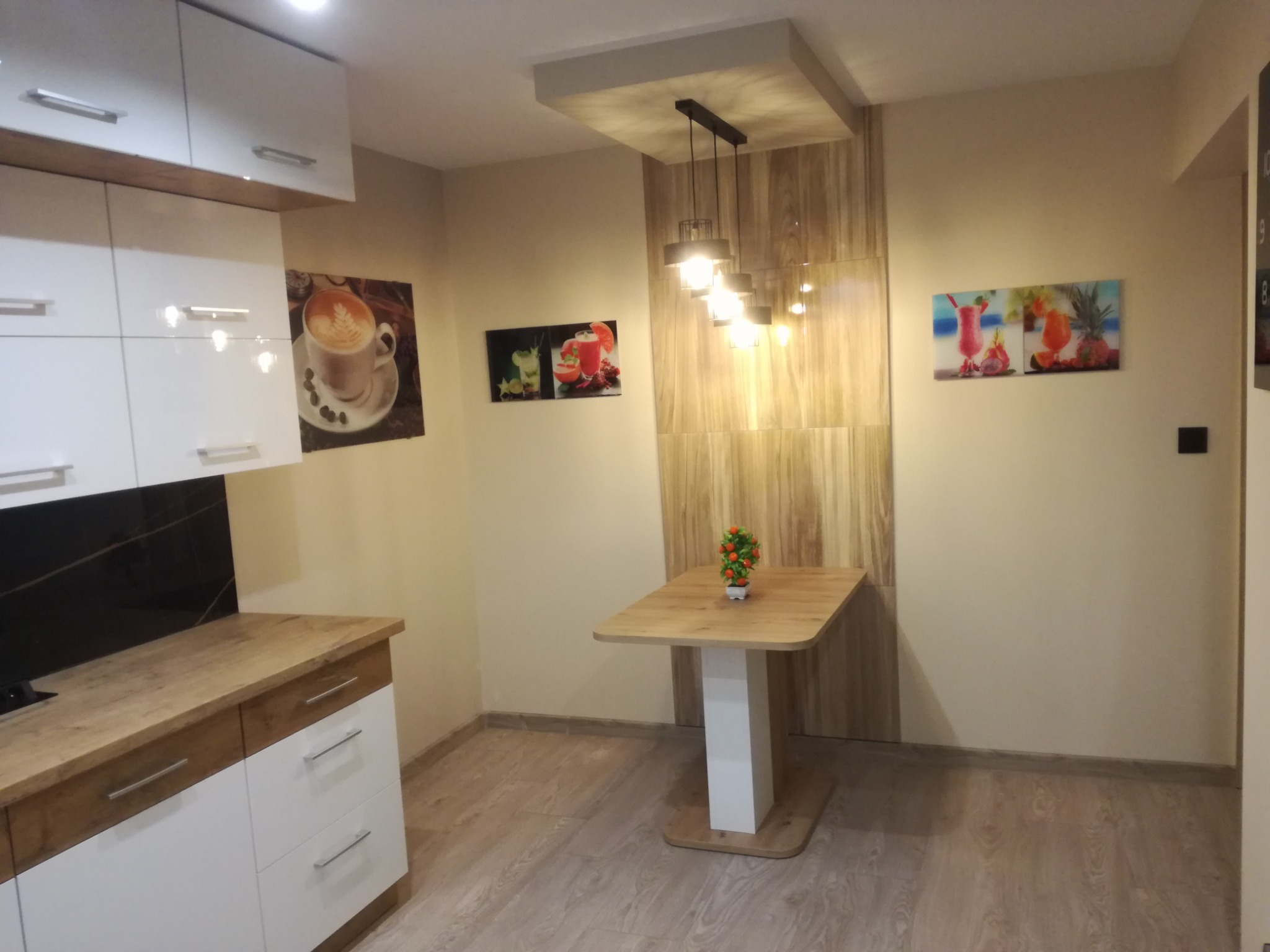 Jasna kuchnia z białymi szafkami, drewnianym blatem i stołem z kwiatami, oświetlona wiszącą lampą z drewnianym panelem dekoracyjnym na ścianie.