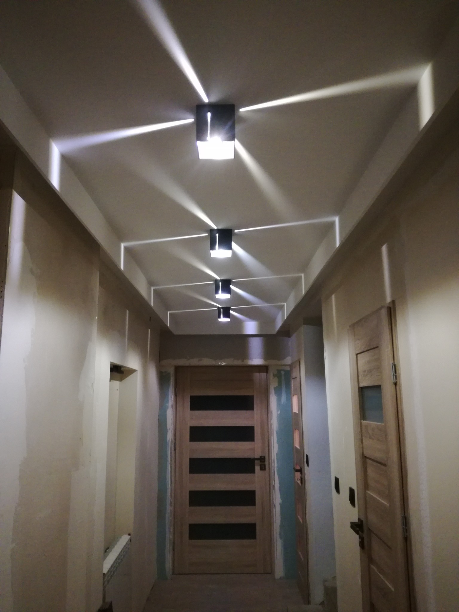 Korytarz w trakcie remontu z zamontowanymi czarnymi lampami sufitowymi, rzucającymi geometryczne światło na sufit i ściany, widoczne ślady po pracach wykończeniowych.