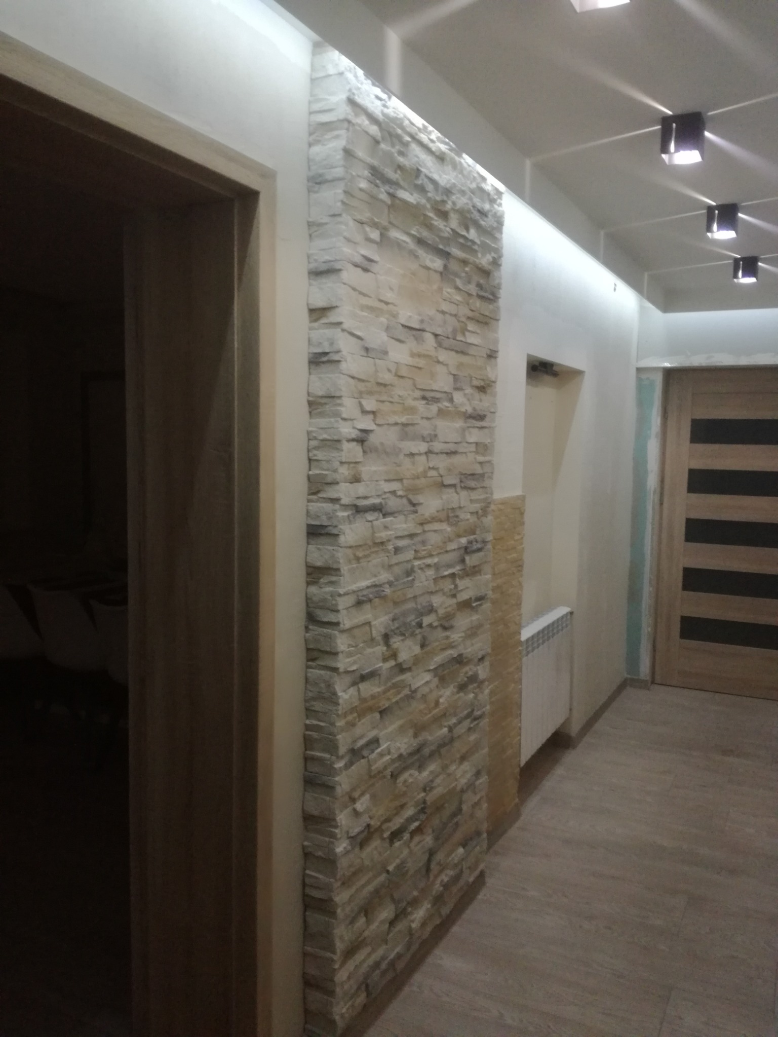 Dekoracyjna ściana z kamienia elewacyjnego w korytarzu, widoczna z perspektywy wejścia do pokoju, z oświetleniem sufitowym i drewnianymi drzwiami w tle.