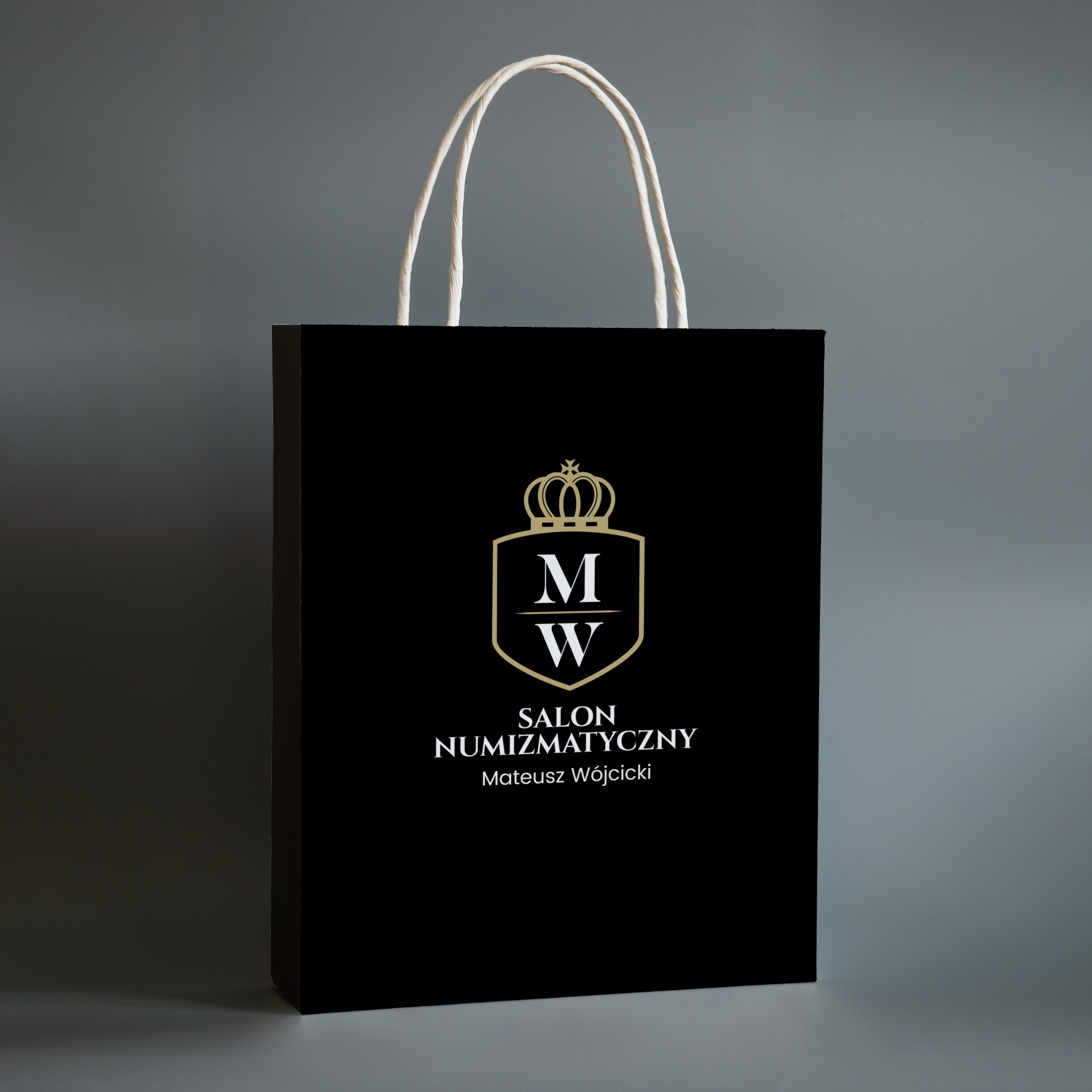 Czarna torba papierowa z białymi uchwytami, na której widnieje złote logo Salonu Numizmatycznego Mateusz Wójcicki.