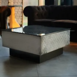 Nowoczesny stolik kawowy z blatem ze spieku kwarcowego i tapicerowanymi bokami w geometryczny wzór, na tle ciemnej sofy i fotela w salonie.