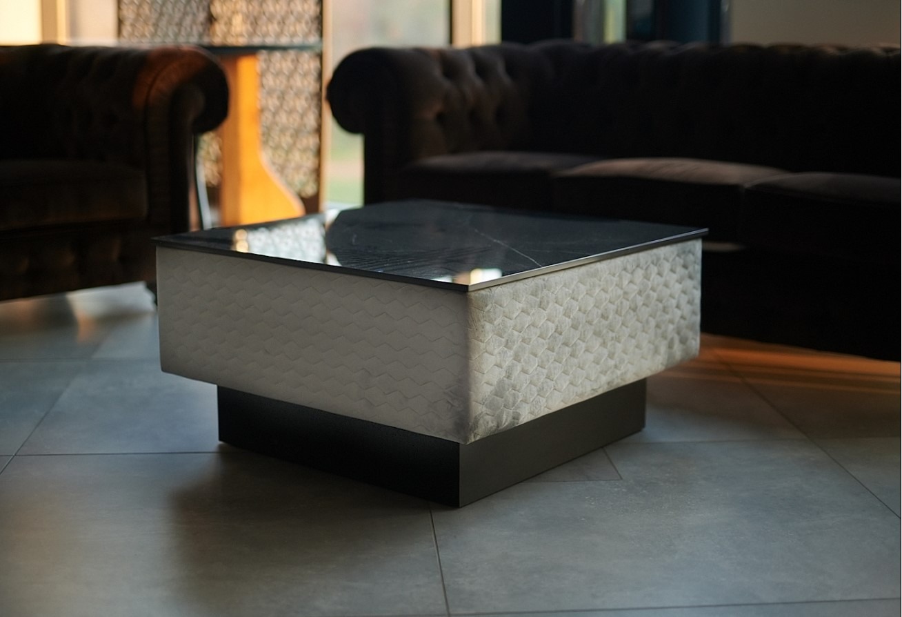 Nowoczesny stolik kawowy z blatem ze spieku kwarcowego i tapicerowanymi bokami w geometryczny wzór, na tle ciemnej sofy i fotela w salonie.
