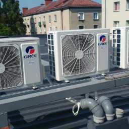 Trzy jednostki zewnętrzne klimatyzacji marki Gree zamontowane na metalowej konstrukcji na dachu budynku mieszkalnego, widoczne rury odprowadzające skropliny.